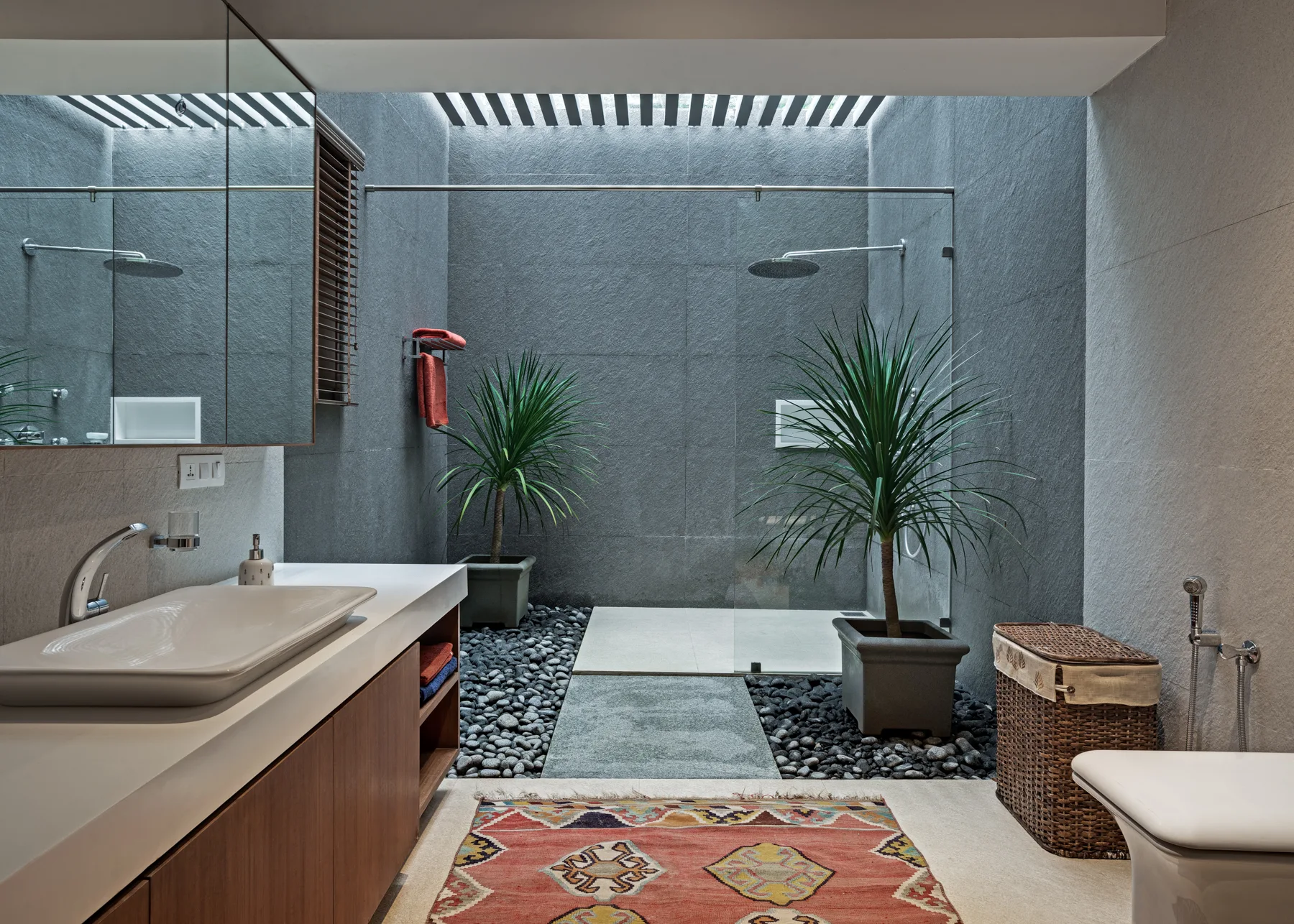 A modern grey stone bathroom features a glass shower stall over a river rock floor, a slatted ceiling skylight, two potted plants, and a wood vanity.