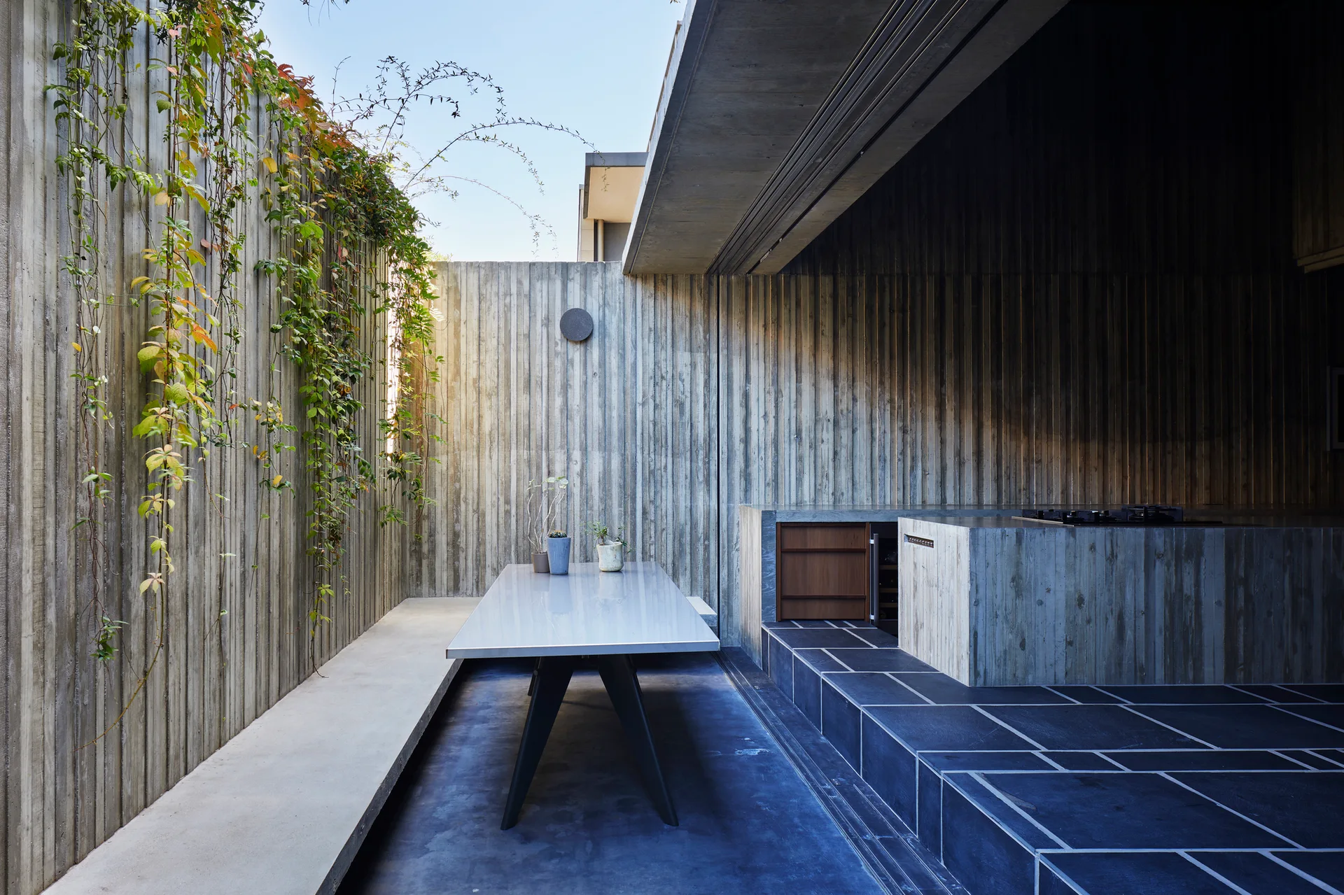 A minimalist outdoor patio features a metal dining table, a built-in concrete kitchen island, and climbing vines on board-formed concrete walls.