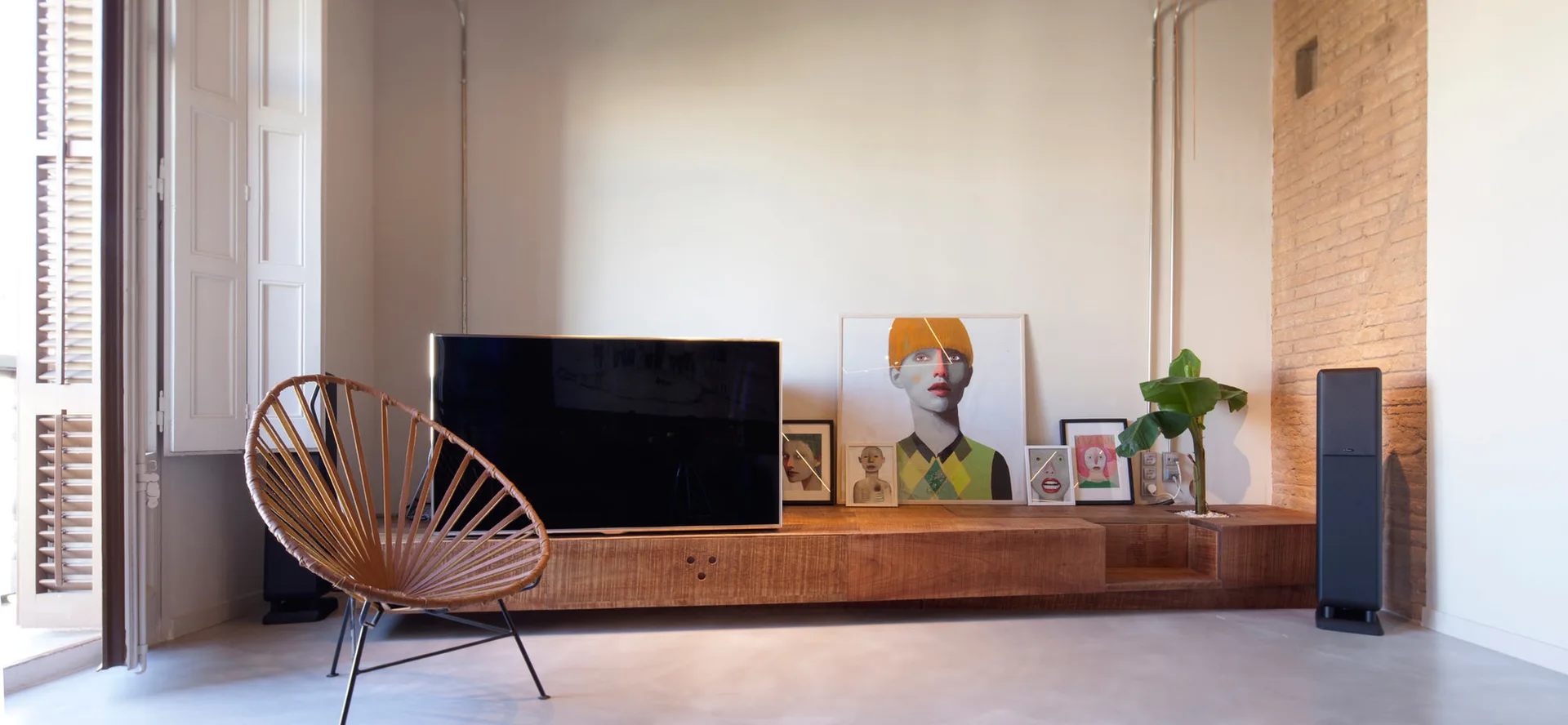 A living room featuring a low wooden media console with a television and leaning framed artwork next to a woven round chair and brick wall.