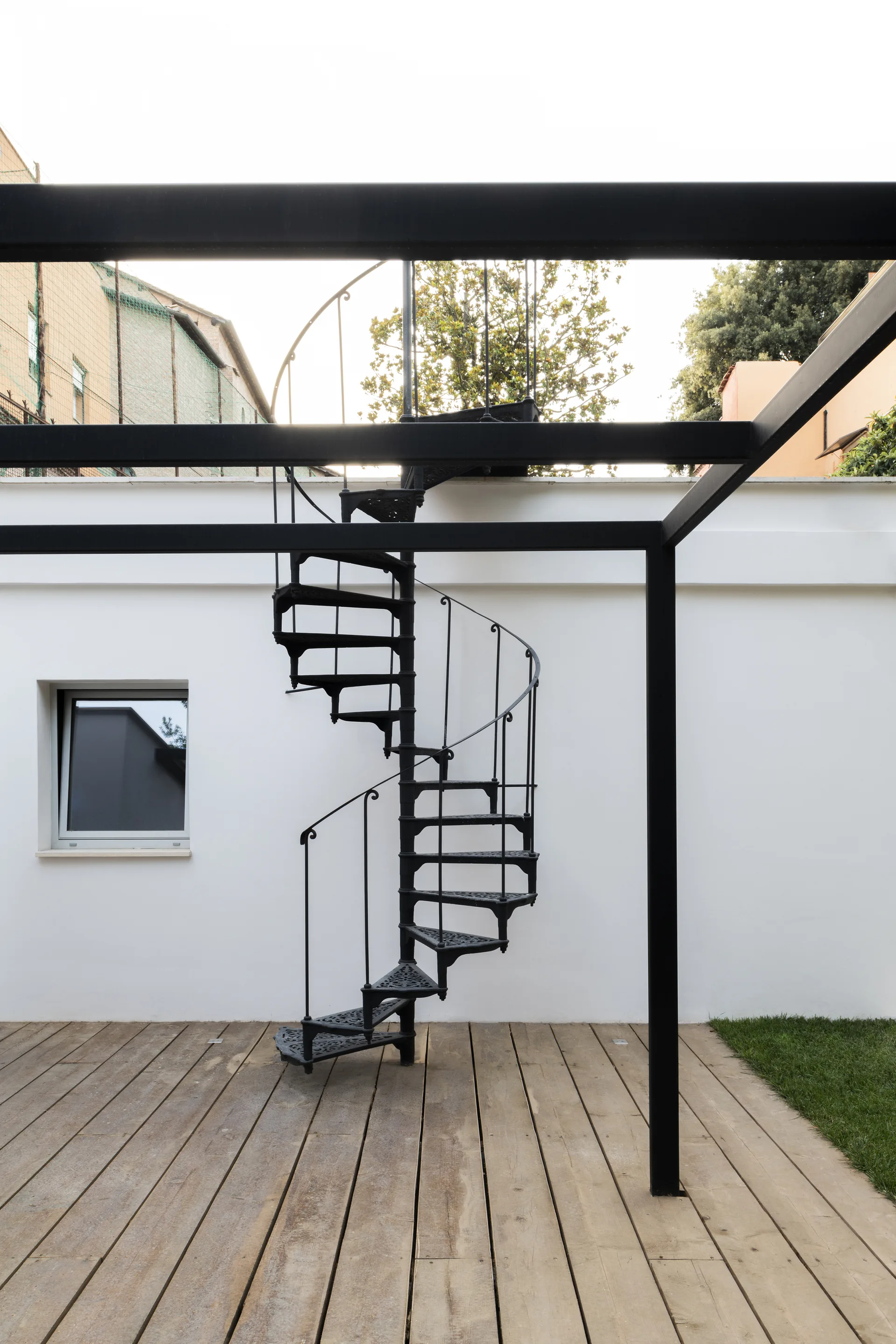 A black metal spiral staircase stands on a light brown wooden deck next to a white stucco wall with a square window under a black overhead frame.