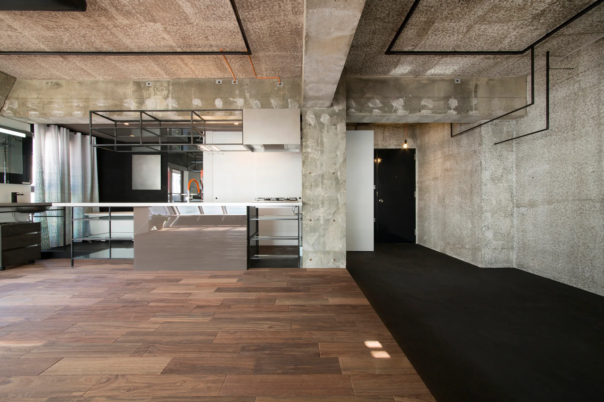 An industrial loft kitchen featuring a minimalist island, raw concrete walls and ceiling, and a sharp split between wood and dark flooring.