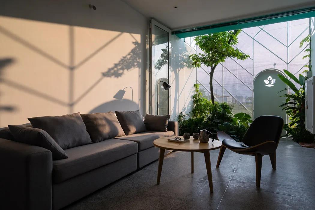 A grey sofa, round wood coffee table, and curved lounge chair sit in a minimalist living room next to a glass wall showing an indoor courtyard.