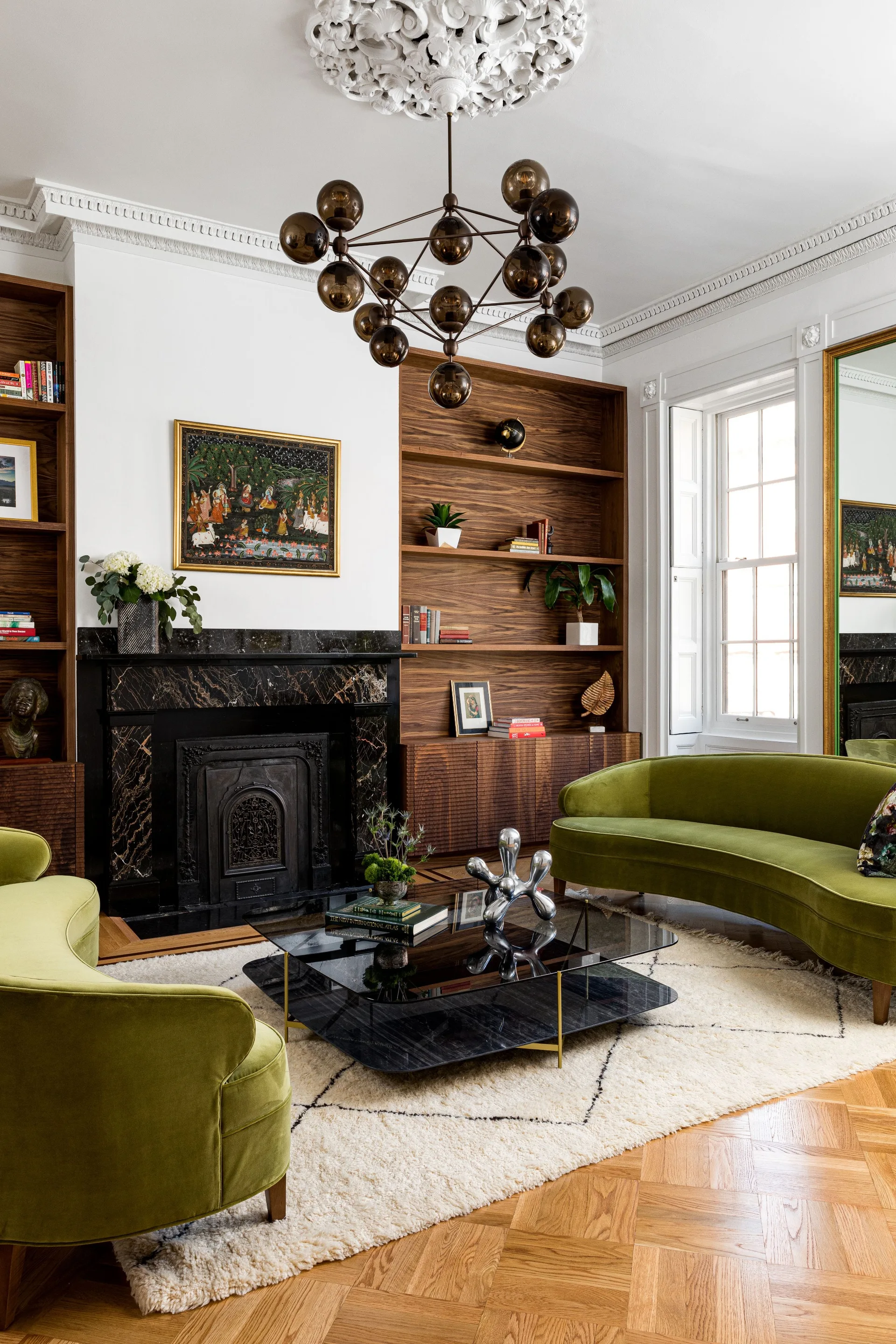 Curved olive green velvet sofas face a black glass coffee table on a shag rug in front of a black marble fireplace and wood built-ins.