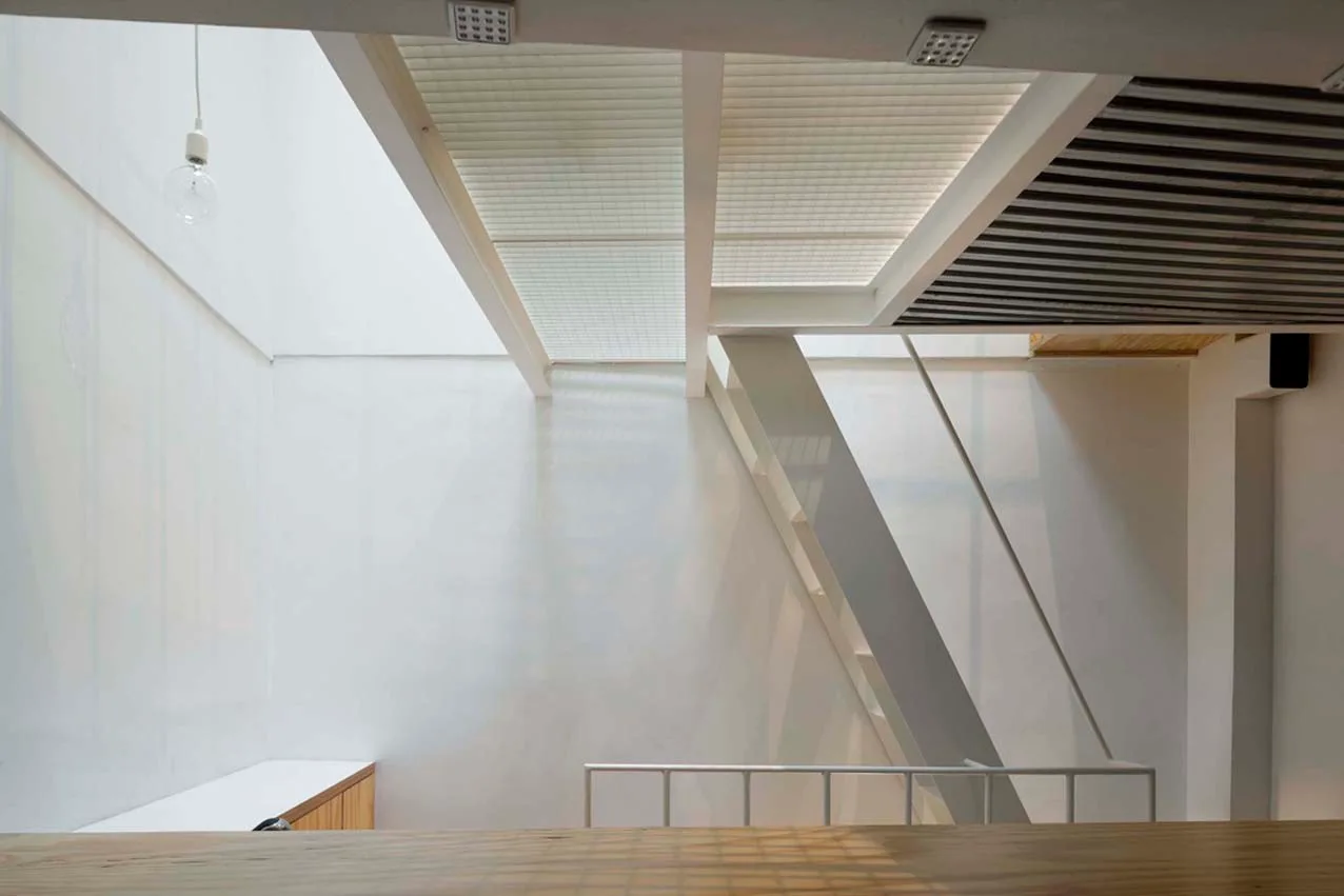 A minimalist white staircase descends from a wooden loft platform under translucent ceiling screens and white beams.