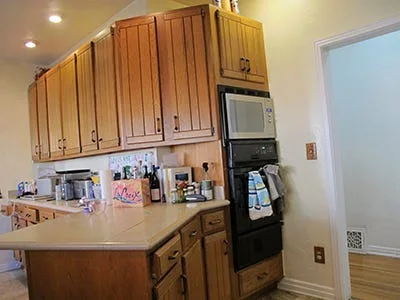A traditional kitchen features vertical-planked wood cabinets, light countertops, and a stacked built-in microwave and black wall oven.
