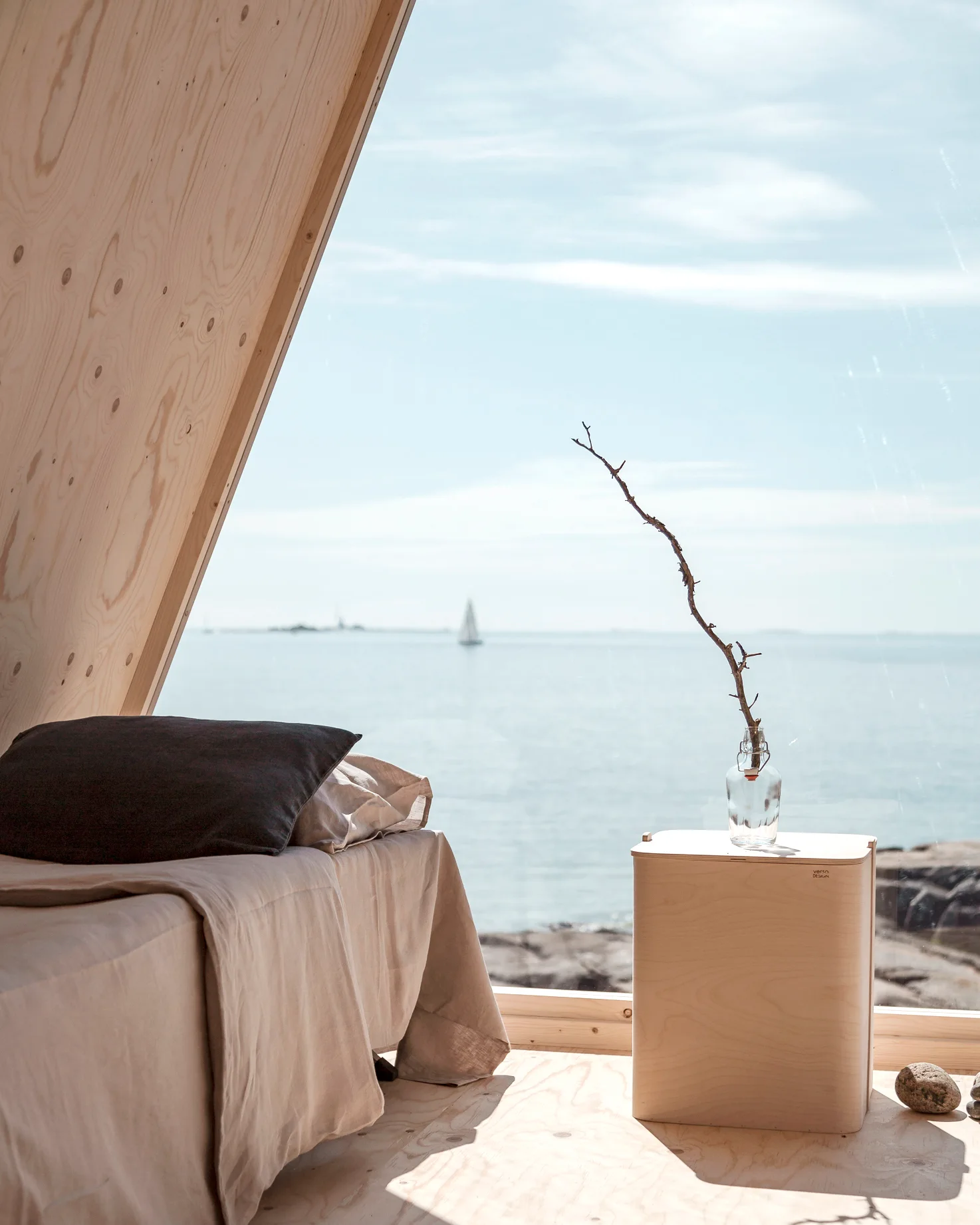 A minimalist cabin bedroom with plywood walls features a bed with beige linens and a small wooden side table next to a large window overlooking the ocean.