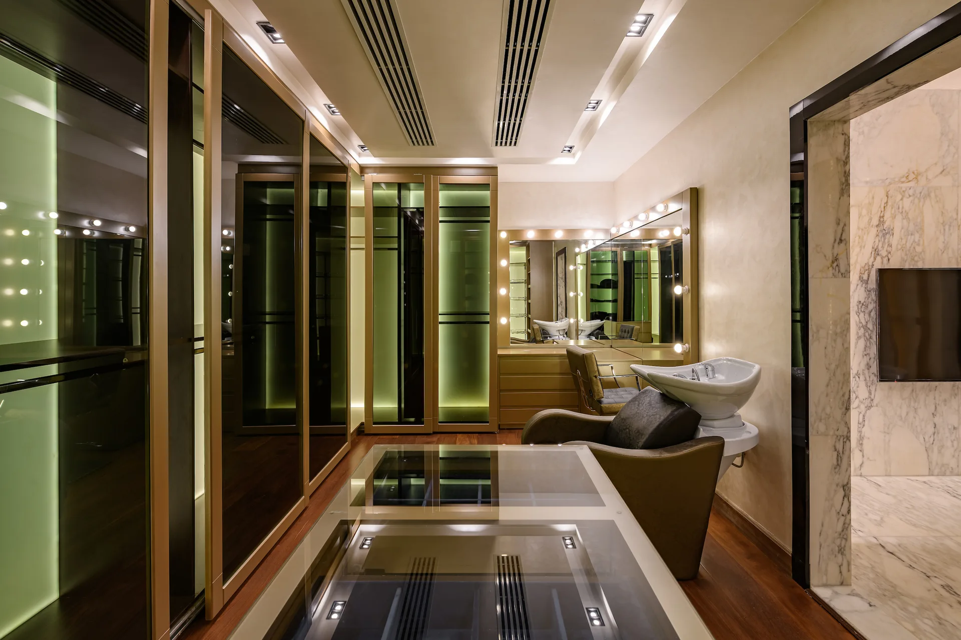 A dressing room features backlit glass wardrobes, a professional salon washing station, a lit vanity mirror, and a glass-top island.