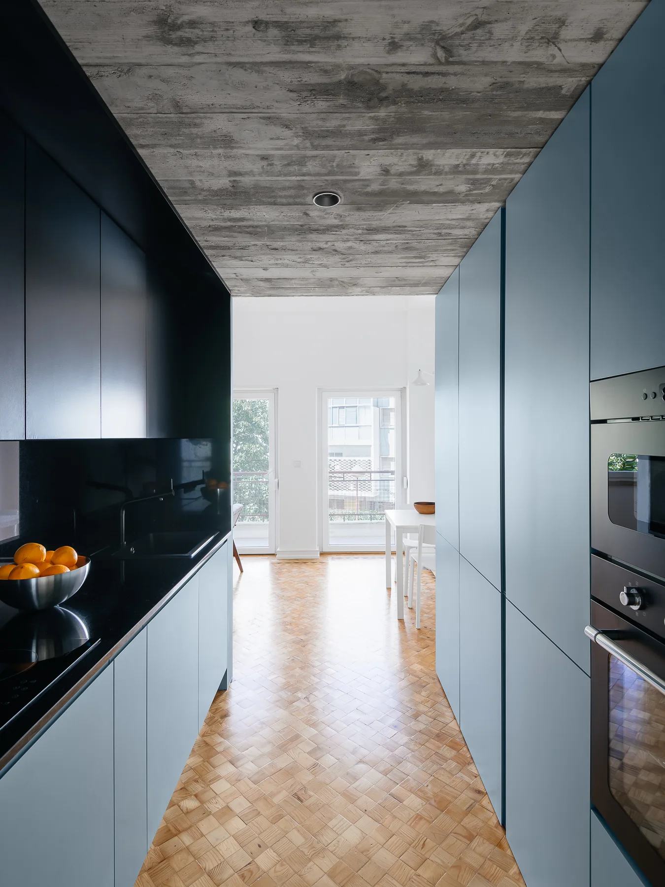 A galley kitchen featuring matte blue and black cabinets, a raw concrete ceiling, and herringbone parquet wood flooring leading to a dining area.