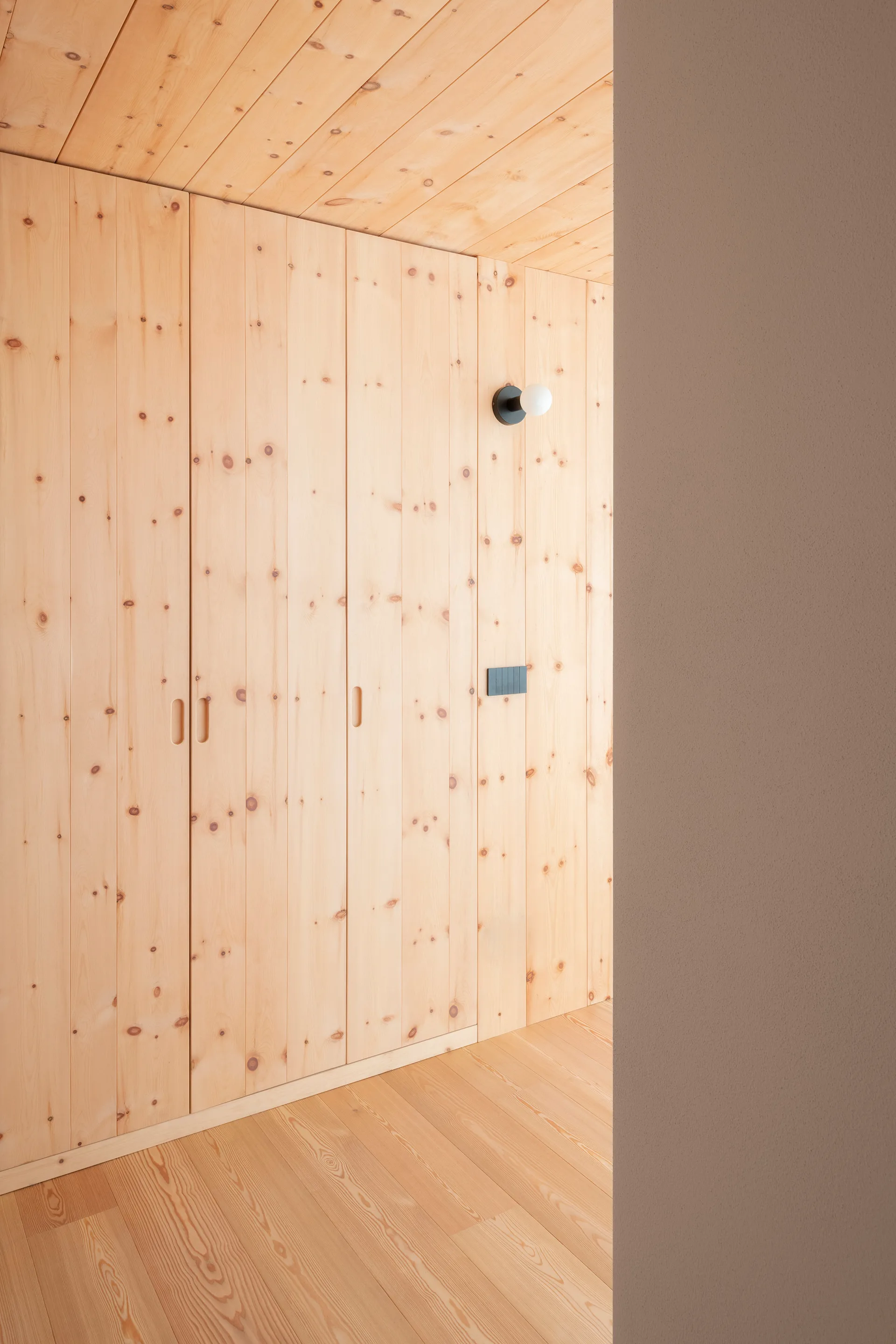 A minimalist hallway lined with floor-to-ceiling knotty pine built-in closets, featuring matching wood ceiling and floors and a black wall sconce.