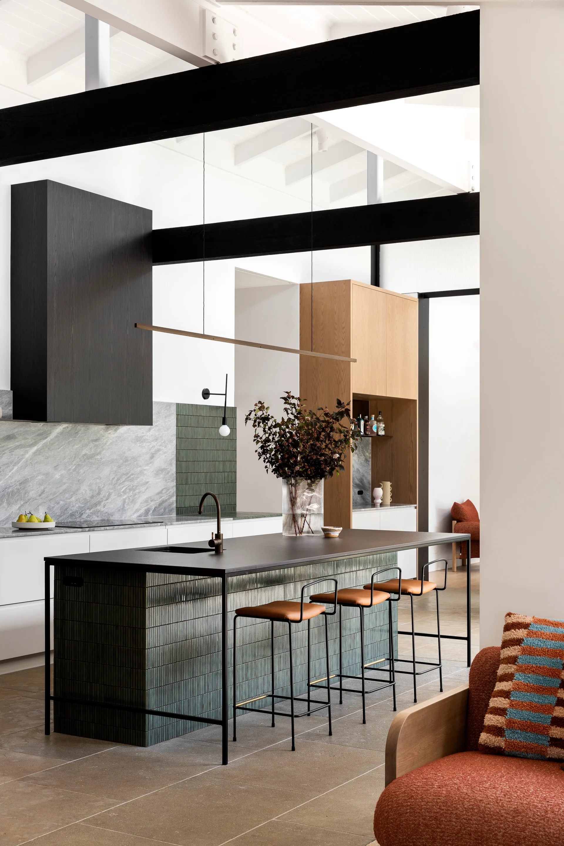 A modern kitchen featuring a black-topped island clad in green fluted tile, leather-seated bar stools, a black range hood, and dark ceiling beams.