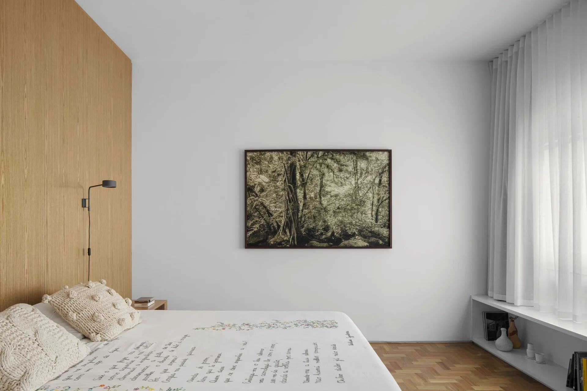 A minimalist bedroom featuring textured throw pillows on a bed, a vertical wood-paneled wall, a large framed nature photograph, and herringbone floors.