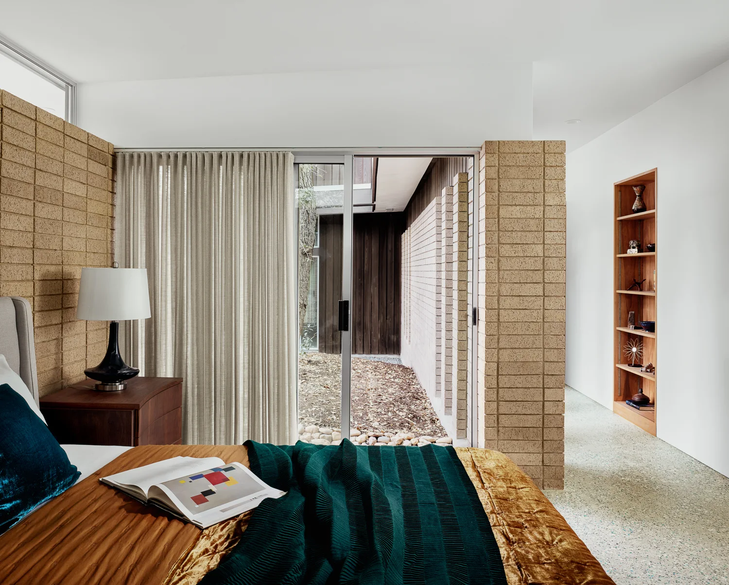 A modern bedroom with copper bedding, a tan brick wall, terrazzo floors, and sliding glass doors that open to an outdoor courtyard.