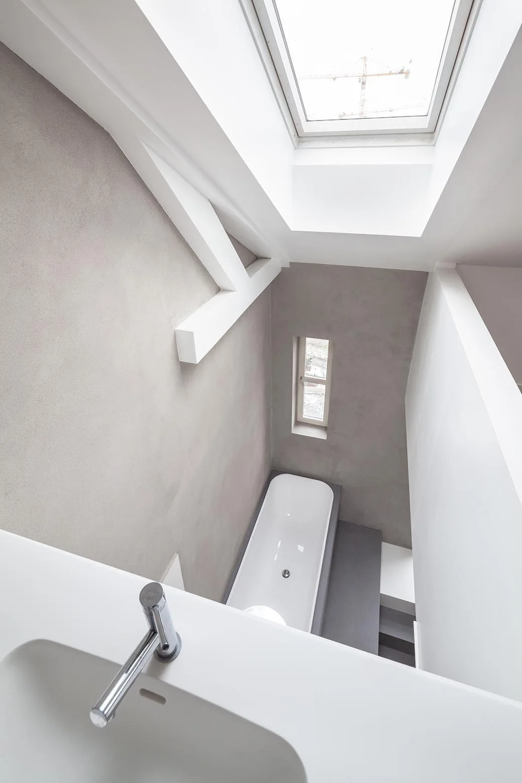 High-angle view of a modern attic bathroom featuring a white bathtub, gray plaster walls, a small window, and a large overhead skylight.