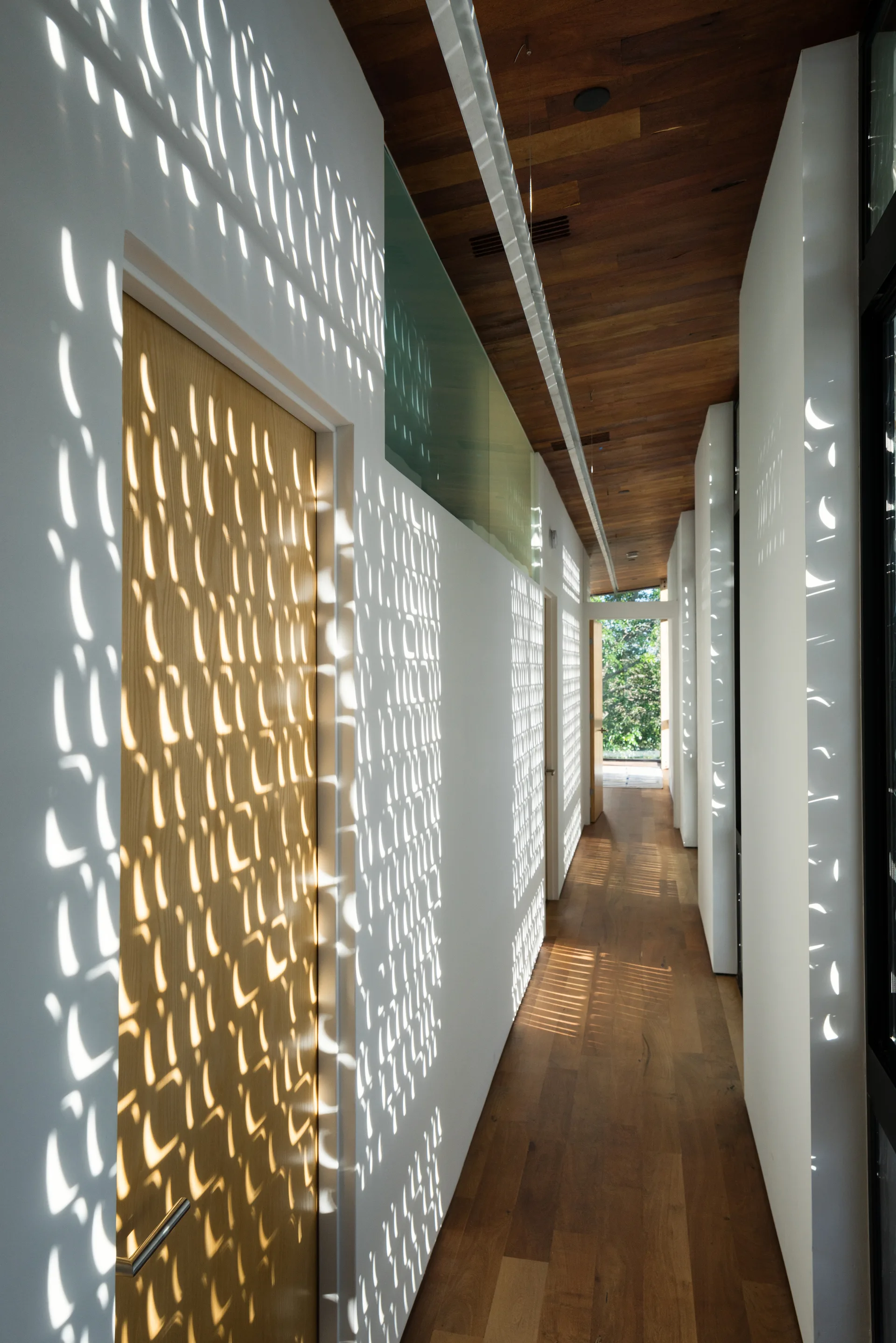 A long hallway features white walls covered in patterned sunlight, a wood plank ceiling, hardwood floors, and a light wood door.