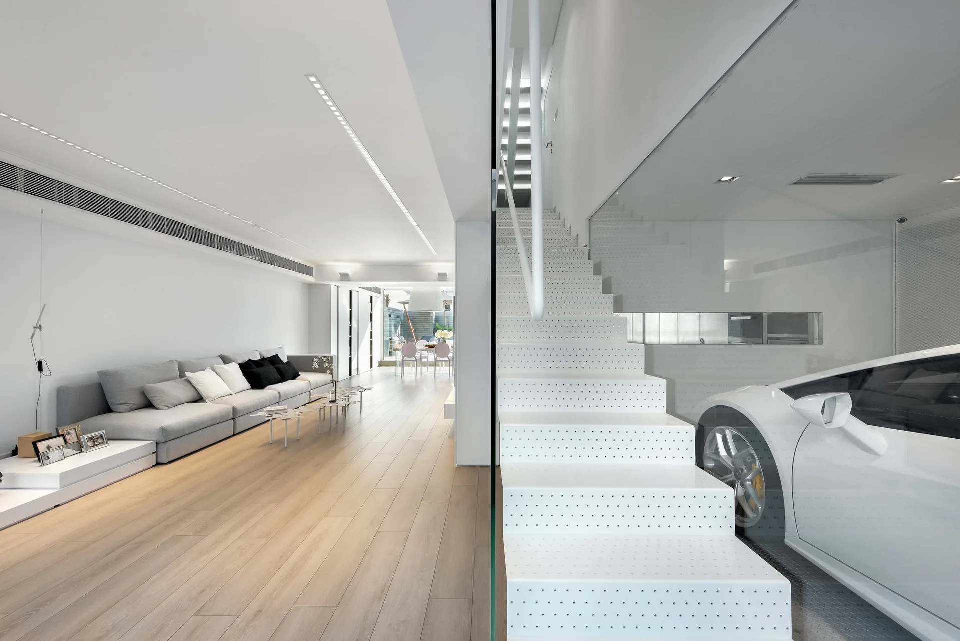 A minimalist living room with a long grey sofa, light wood floors, and a white perforated metal staircase next to a glass-walled garage.