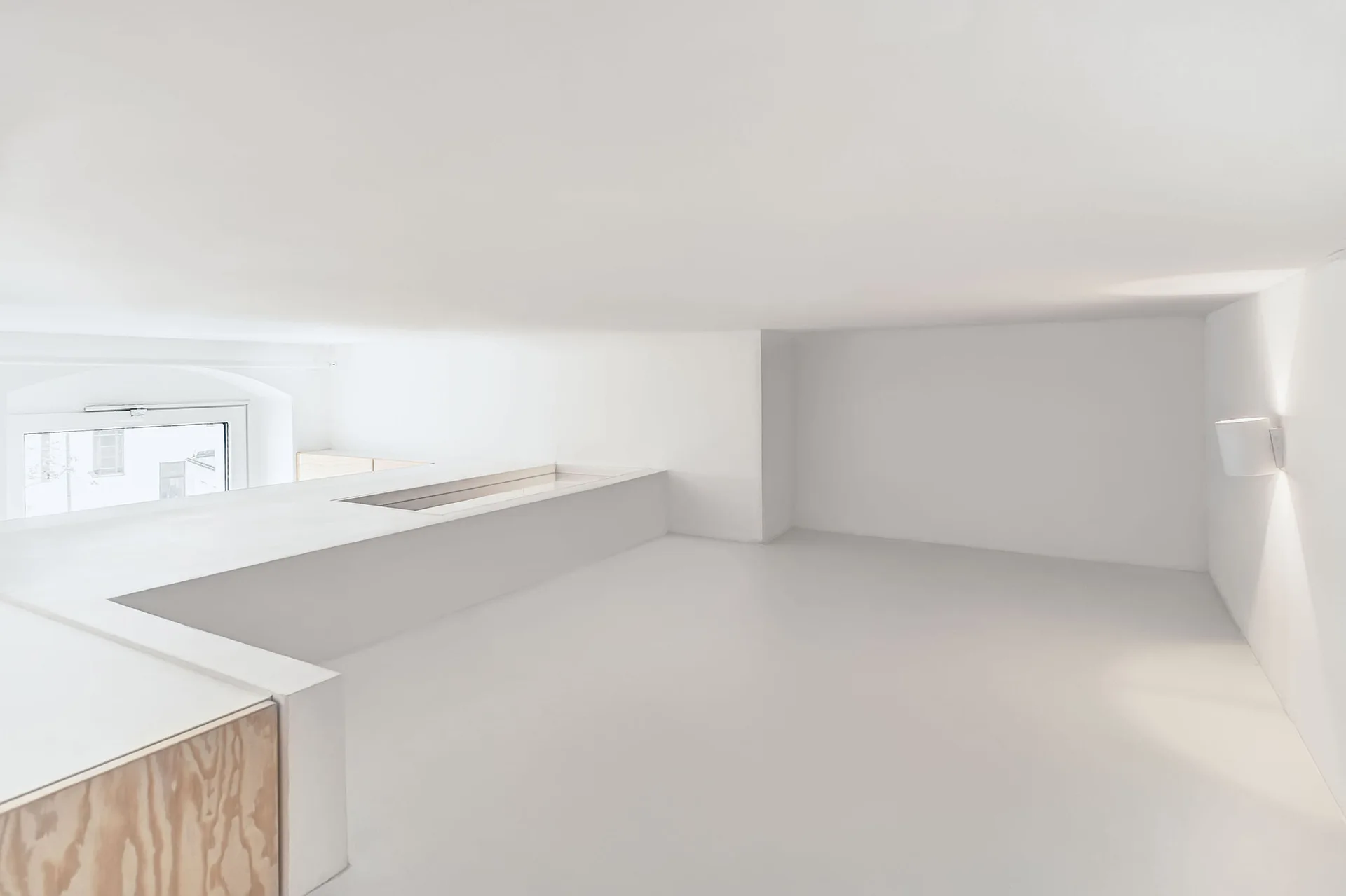 A minimalist white loft bedroom features a low-profile platform bed, a small window, and a wall sconce on a plain white wall.