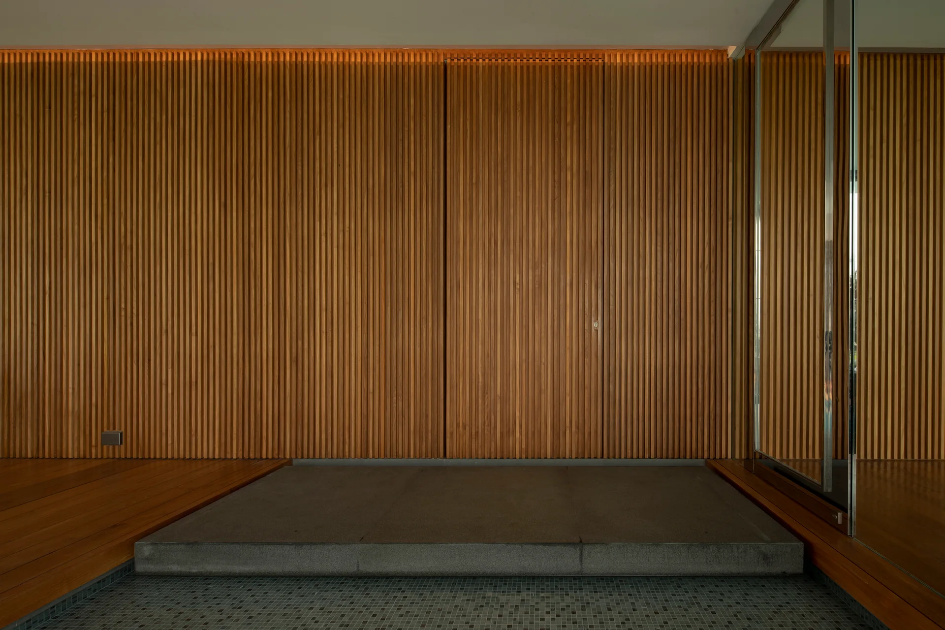 A minimalist wall features vertical wood slats with a flush integrated door above a raised grey stone platform.