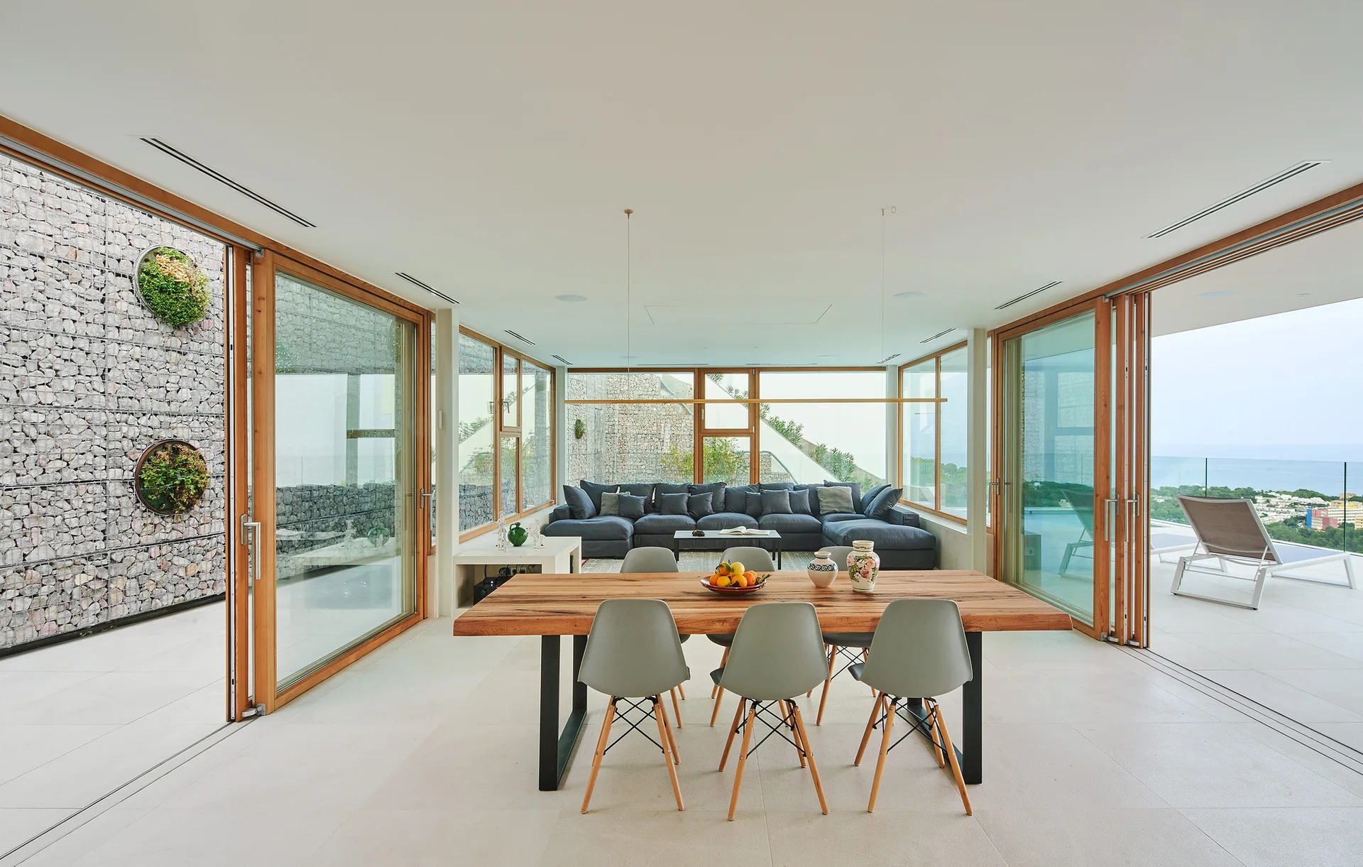 An open-concept room features a wood dining table with grey chairs, leading to a blue sectional sofa and sliding glass doors that open to the outdoors.