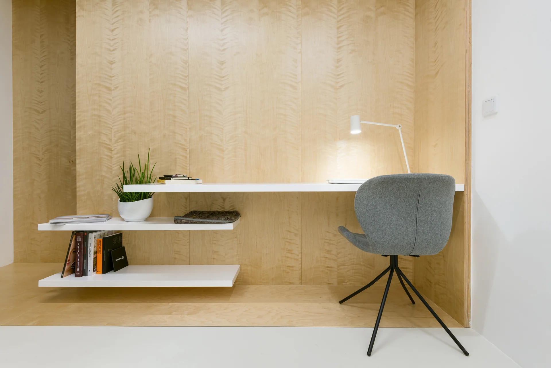 A minimalist home office features a floating white desk and staggered shelves mounted against a light wood-paneled wall, paired with a grey swivel chair.
