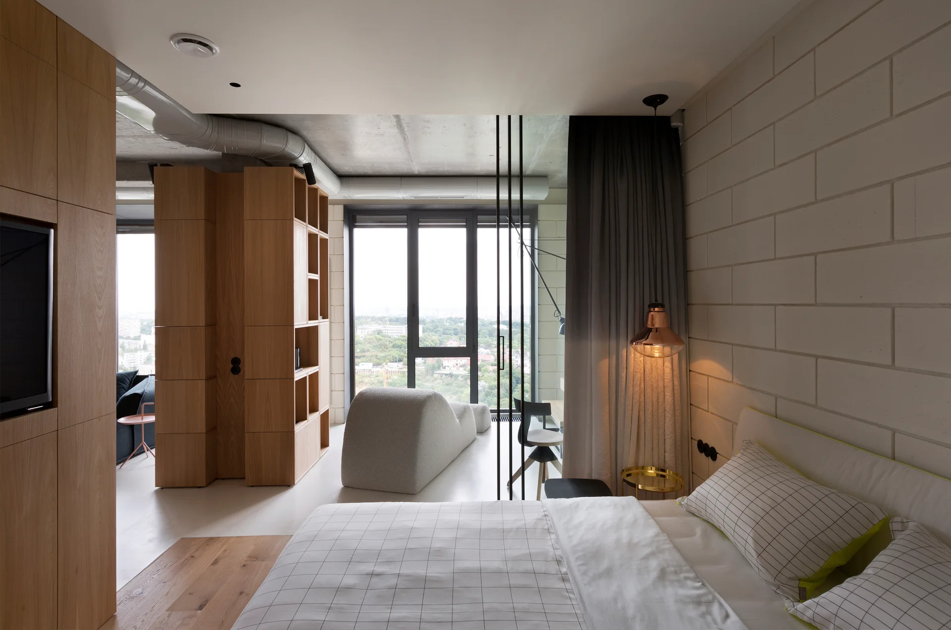 A modern loft bedroom featuring an exposed concrete ceiling, a painted block wall, a large wooden shelving unit divider, and a bed with grid-patterned pillows.