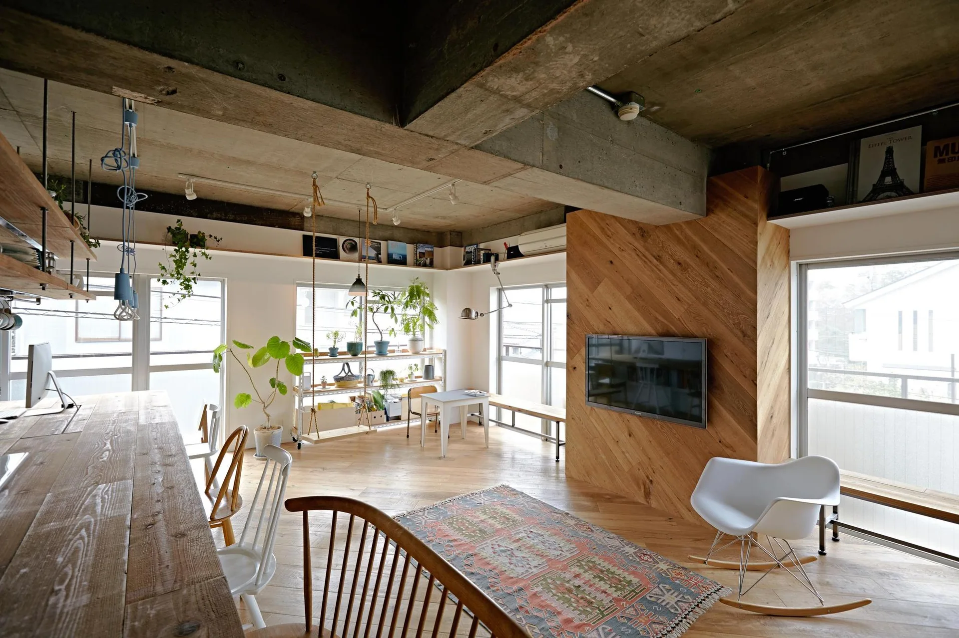 An open-concept living space features exposed concrete ceiling beams, a diagonal wood paneled media wall, and light wood flooring.