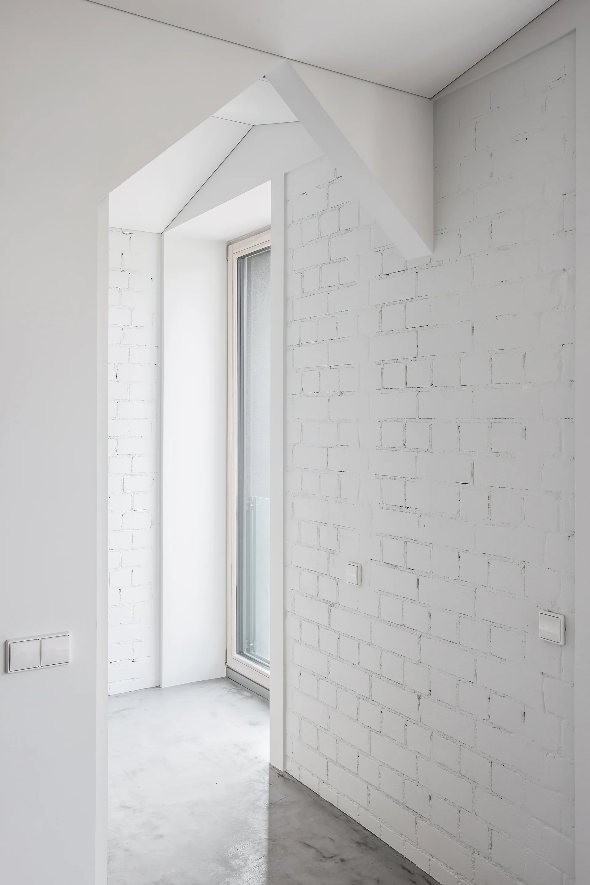 A minimalist hallway featuring white painted brick walls, a polished concrete floor, and a tall narrow window in the background.