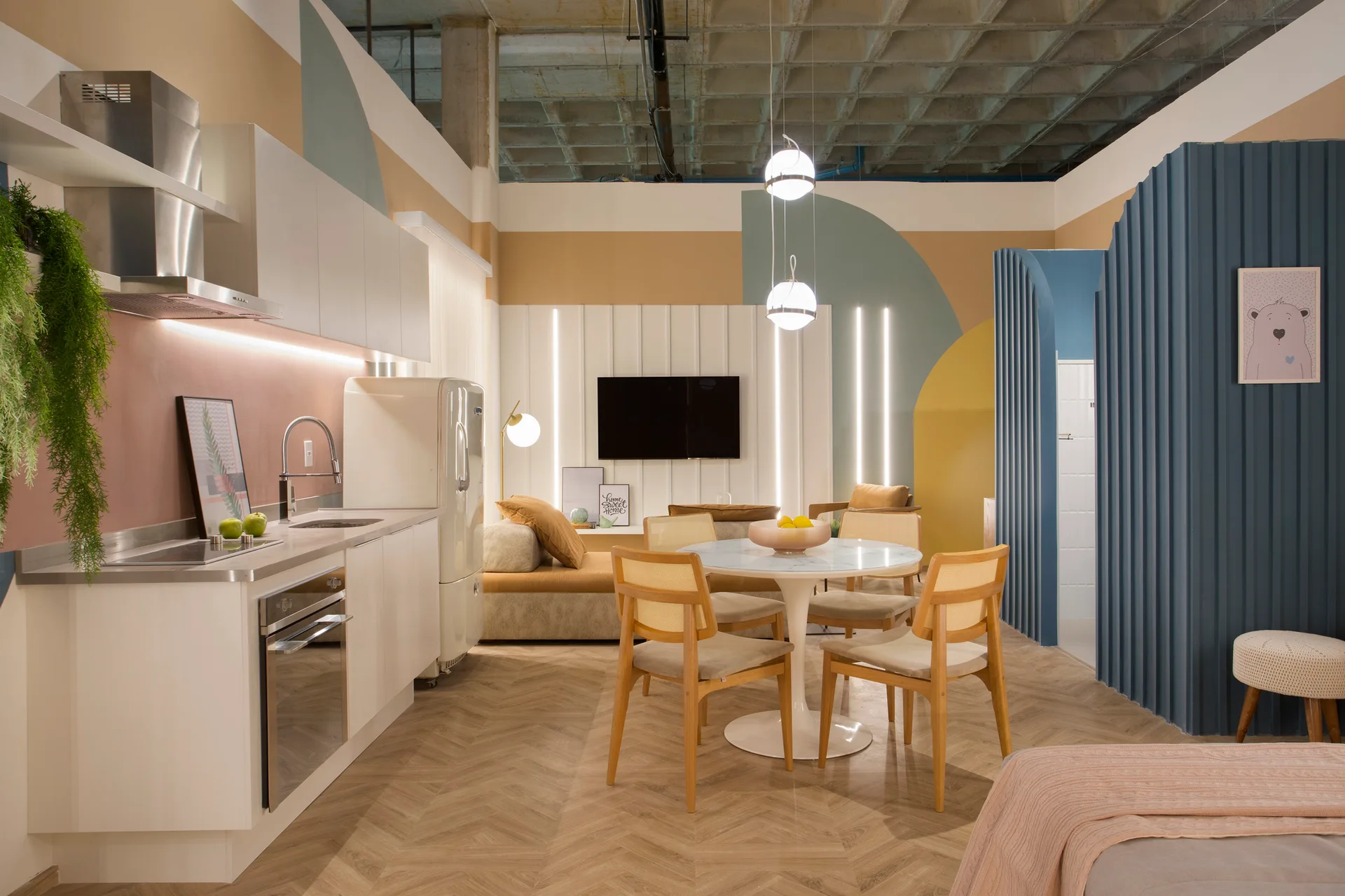An open-concept studio apartment featuring a white kitchen, a dining set with cane chairs, and walls painted with colorful geometric shapes.