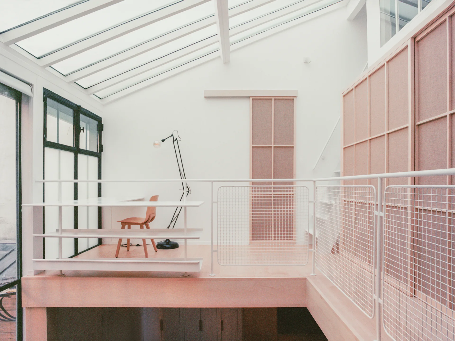 A bright mezzanine office featuring a white desk, wooden chair, and mesh railing beneath a sloped glass roof with paneled wooden cabinets.
