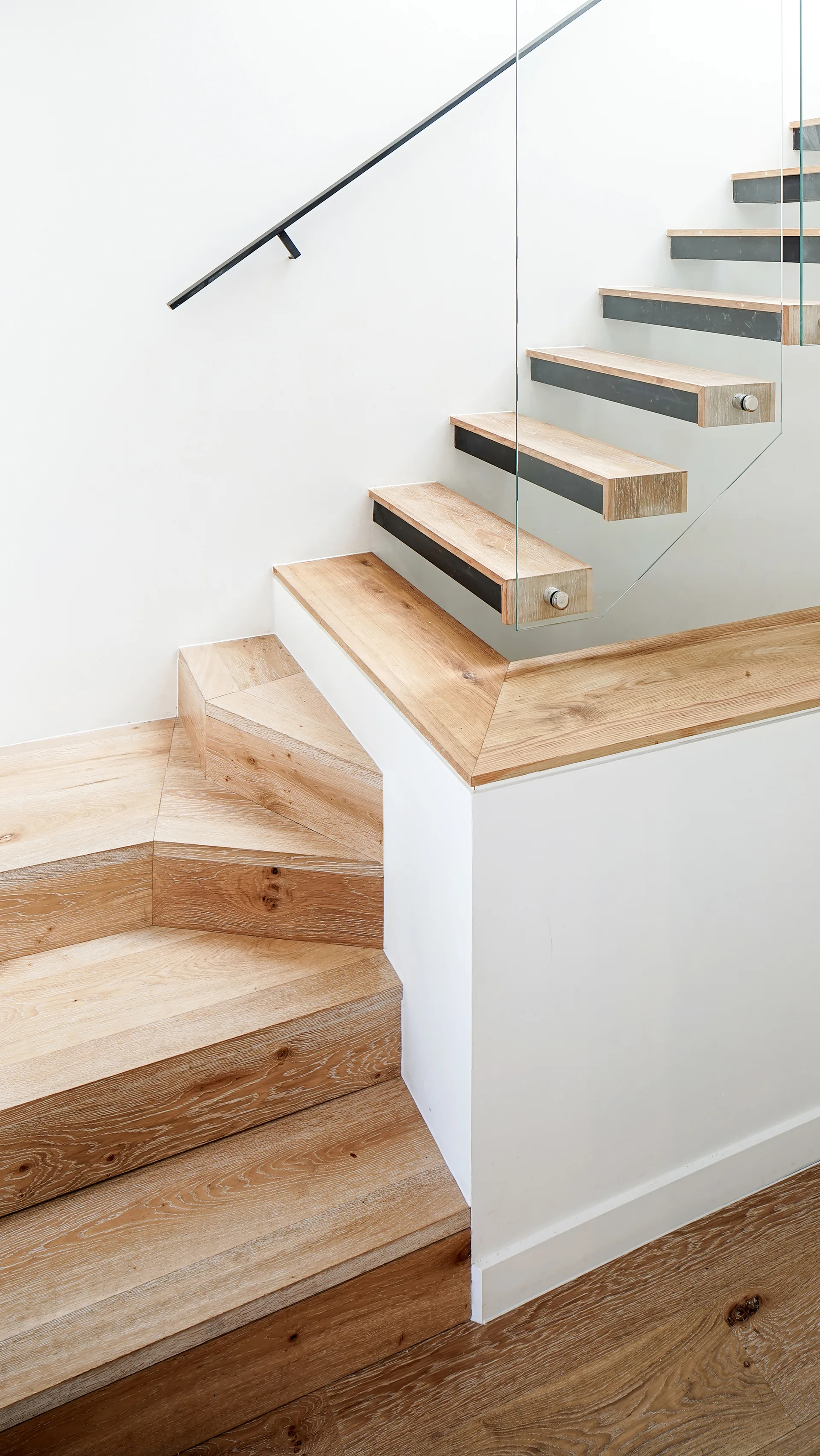 A modern staircase with light-toned wooden treads, a clear glass panel railing, and a simple black metal handrail against a white wall.