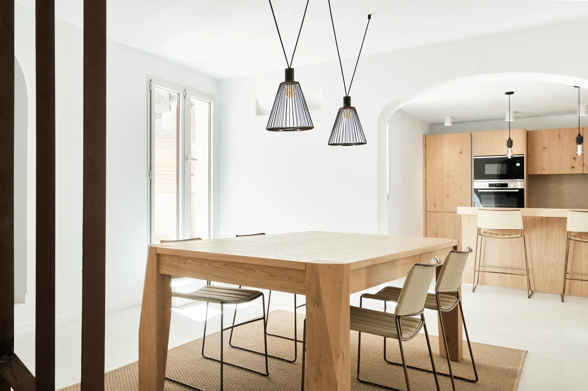 A minimalist dining room features a light wood table, black wire pendant lights, and an arched doorway leading to a kitchen with wood cabinets.