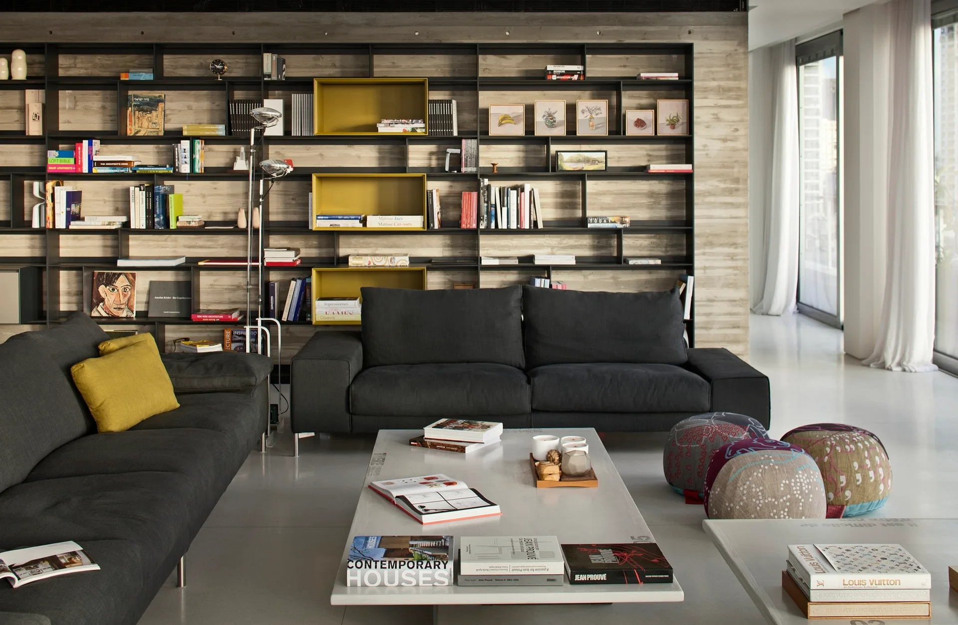 A modern living room with a large black metal shelving unit on an exposed concrete wall, two dark gray sofas, and a white coffee table.