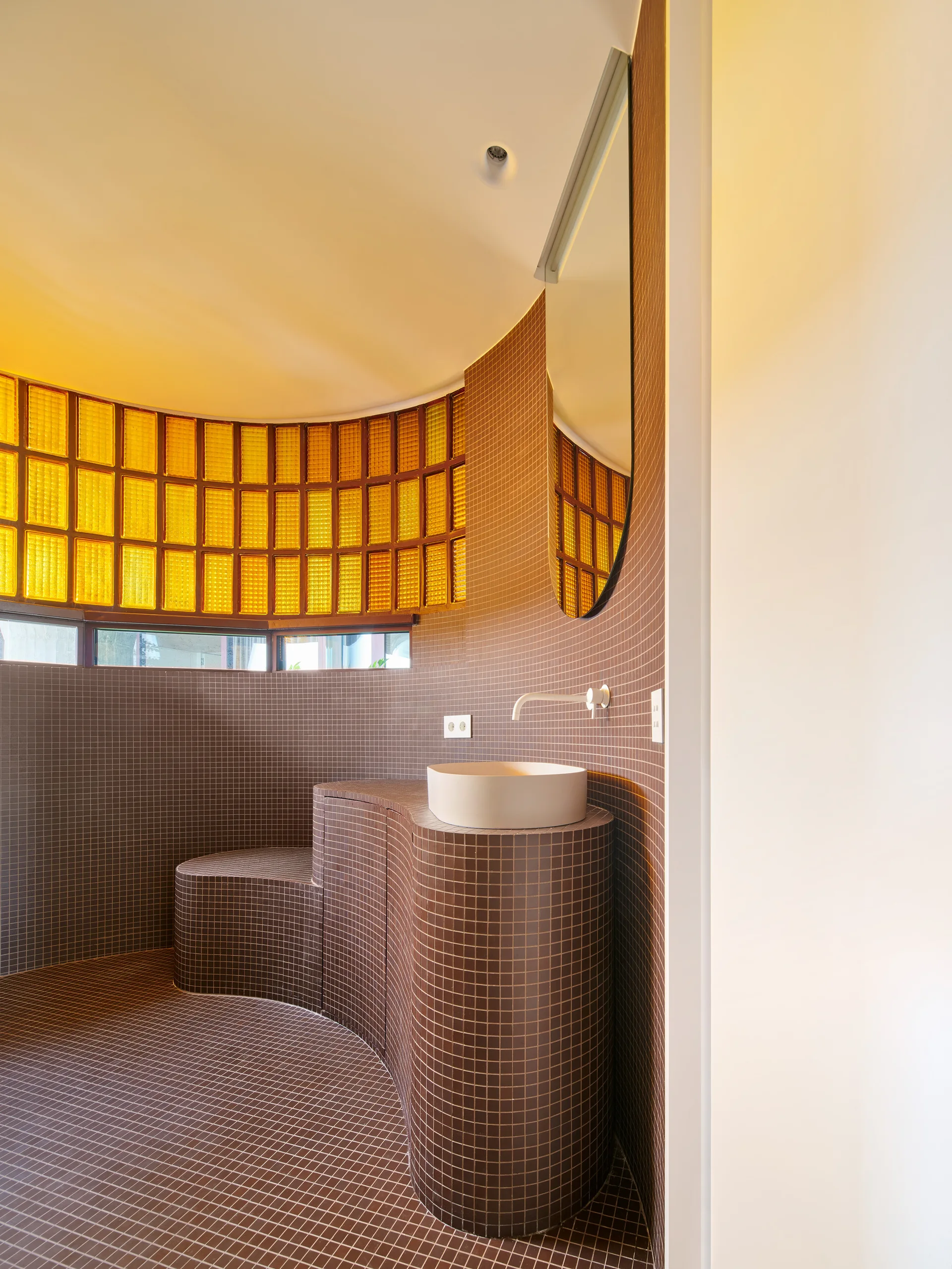 A modern bathroom features a curved vanity clad in brown mosaic tiles, a round vessel sink, and a yellow glass block window.