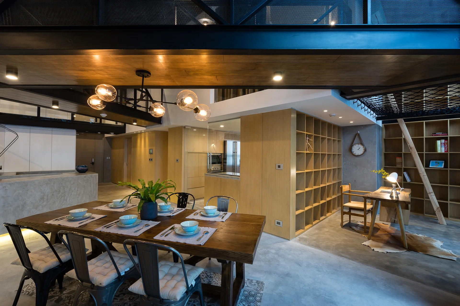 An industrial loft dining room features a dark wood dining table with metal chairs, a light wood grid bookshelf, and heavy black steel ceiling beams.