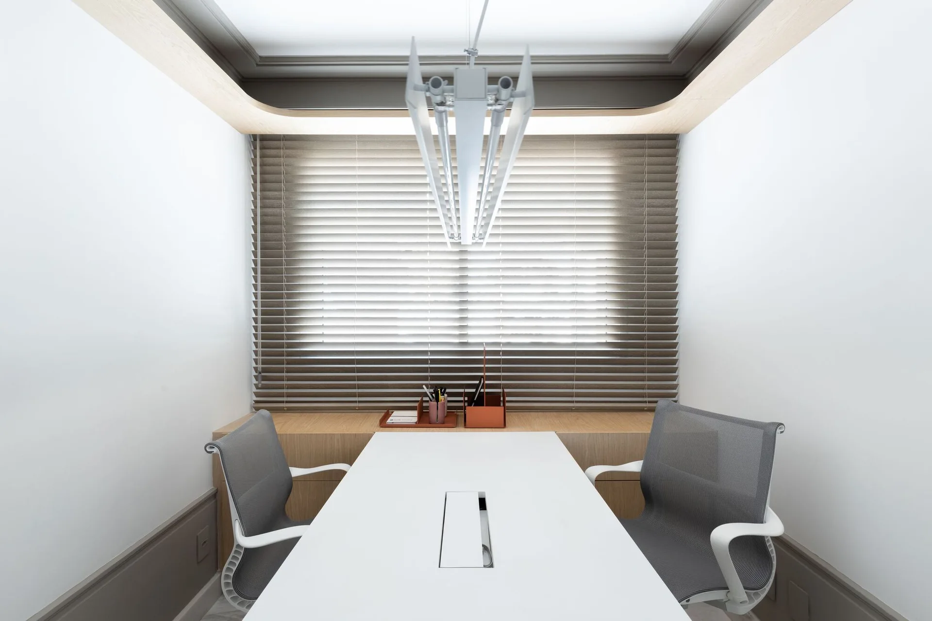 A minimalist home office features a central white desk flanked by grey mesh chairs in front of a window with slatted blinds and wood cabinets.