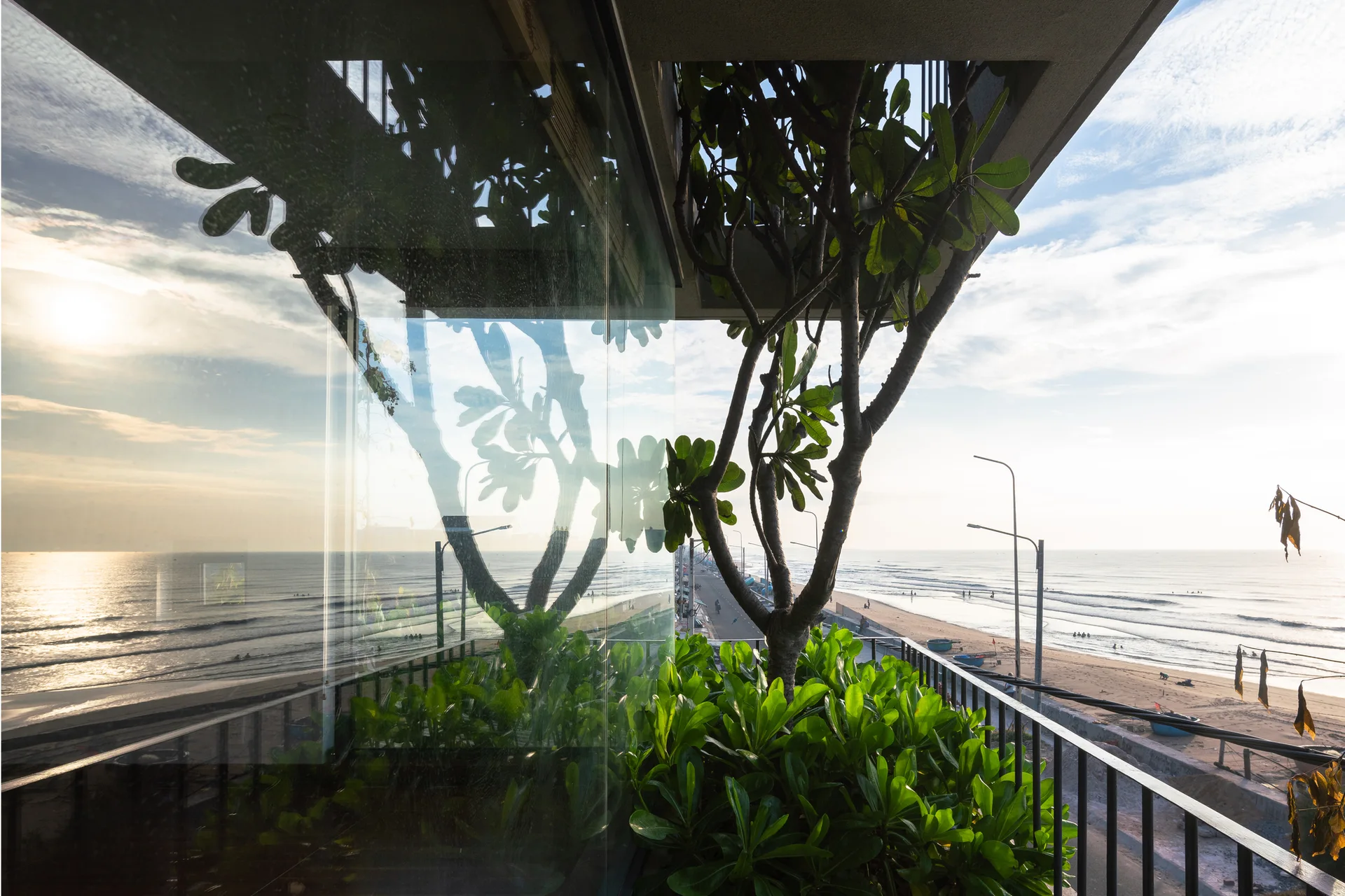 An outdoor balcony with a black metal railing, a large reflective glass panel, and lush green plants overlooking a sandy beach and ocean at sunset.