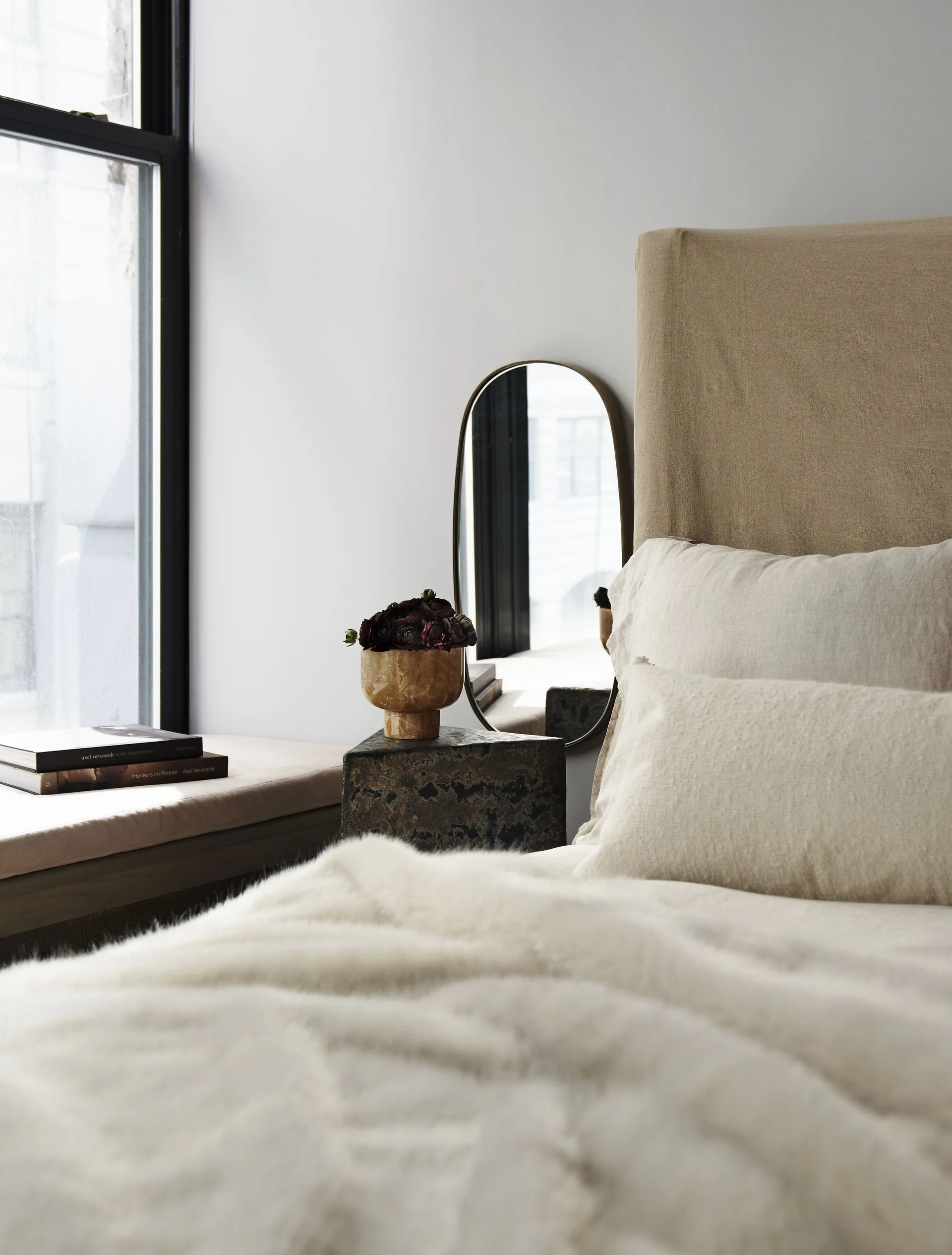 A minimalist bedroom features a beige linen upholstered headboard, a dark textured stone nightstand with a floral arrangement, and a slim oval mirror.