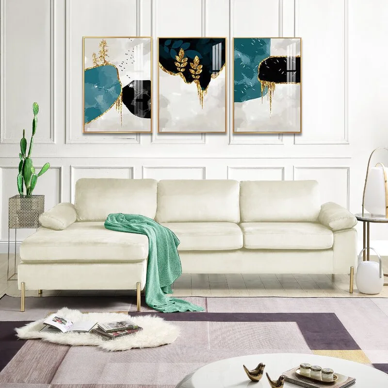 A cream velvet sectional sofa sits in front of a white paneled wall decorated with three abstract teal and gold art panels.