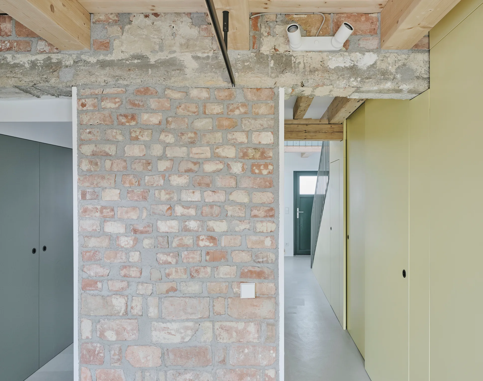 A modern residential hallway featuring an exposed red brick wall, pale yellow flat-panel cabinets, exposed wood ceiling beams, and concrete floors.