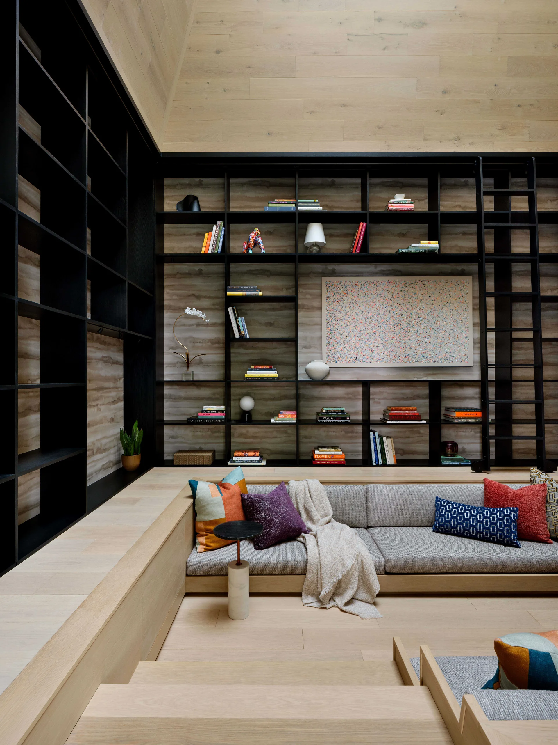 A sunken living room conversation pit features a gray sofa next to light wood platforms and a floor-to-ceiling black bookcase with a library ladder.