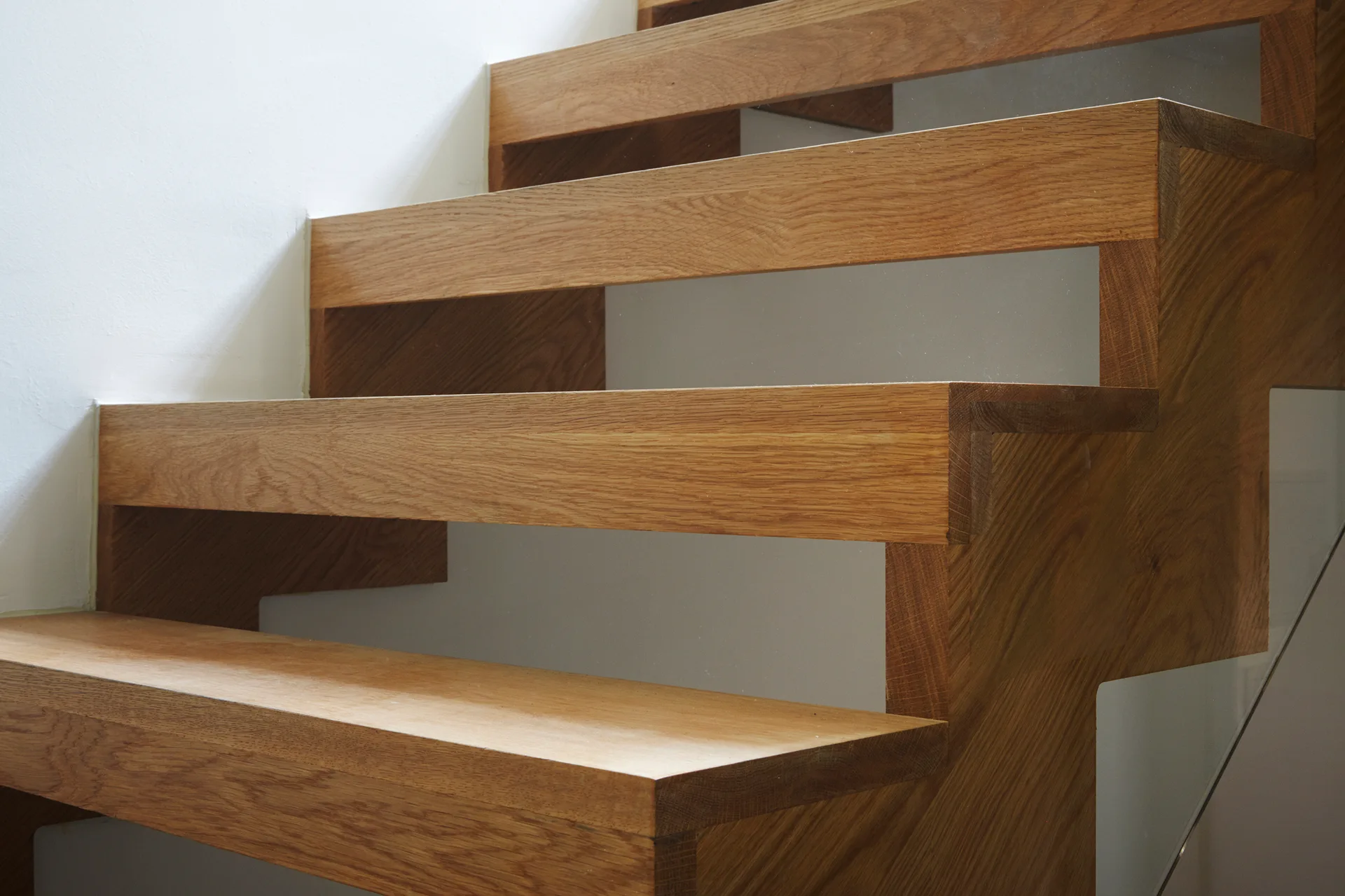 Modern wood staircase with thick stepped timber treads and a glass balustrade set against a plain white painted wall.