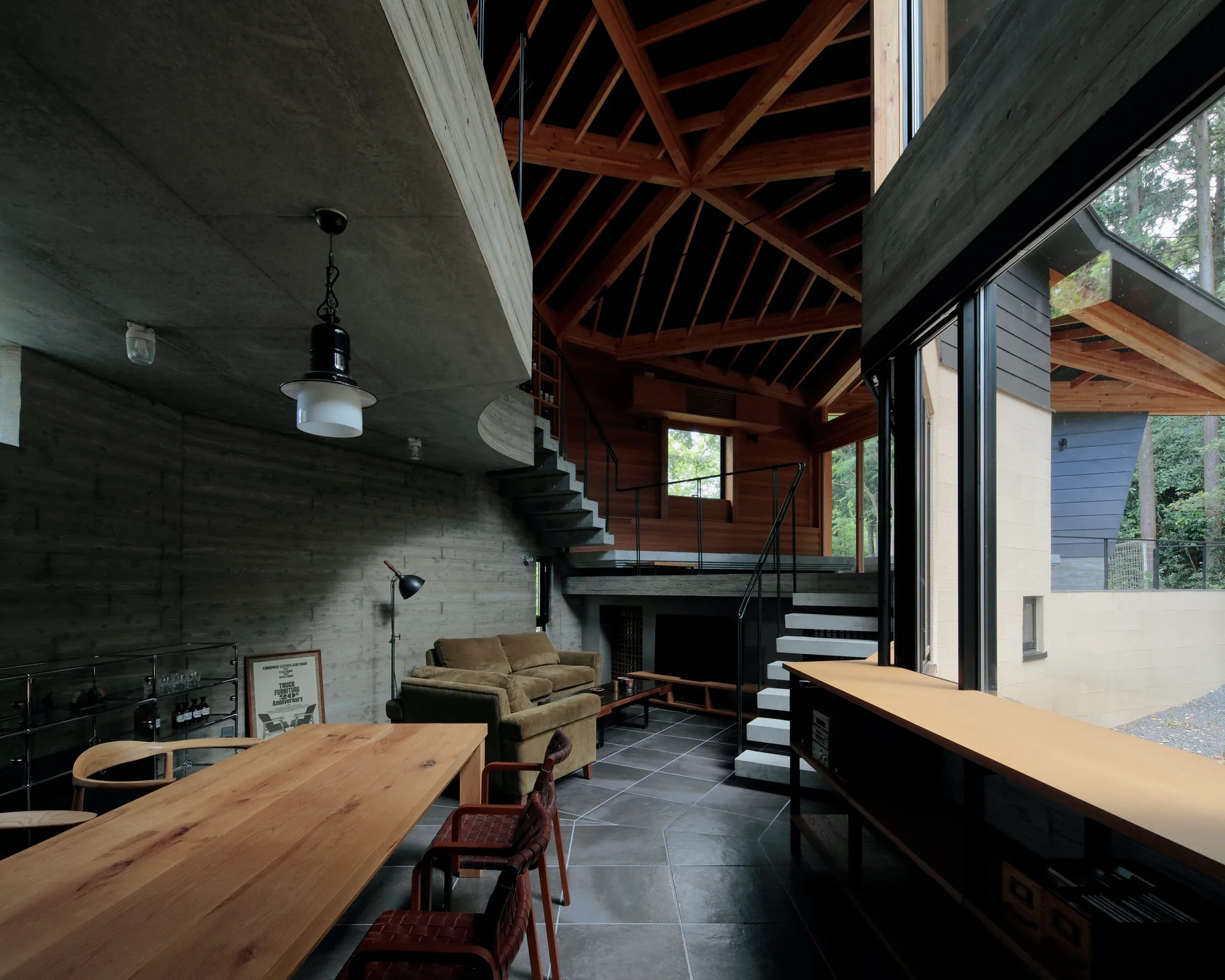 A modern living space features a complex timber truss ceiling, board-formed concrete walls, and floating concrete stairs leading to a split-level area.