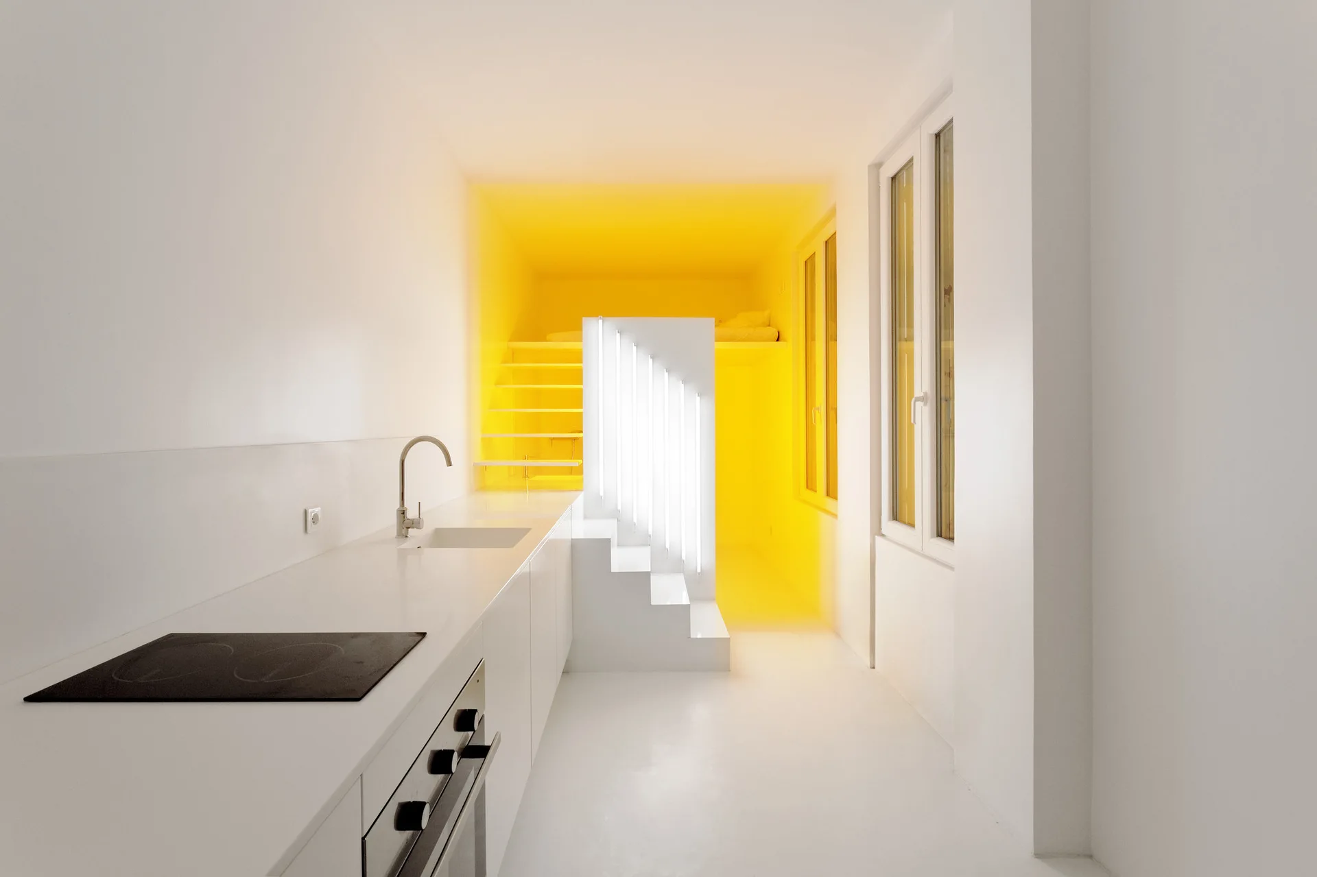A minimalist white kitchen features a long counter with a cooktop, a tall window, and a white staircase leading to a vibrant yellow lit mezzanine area.