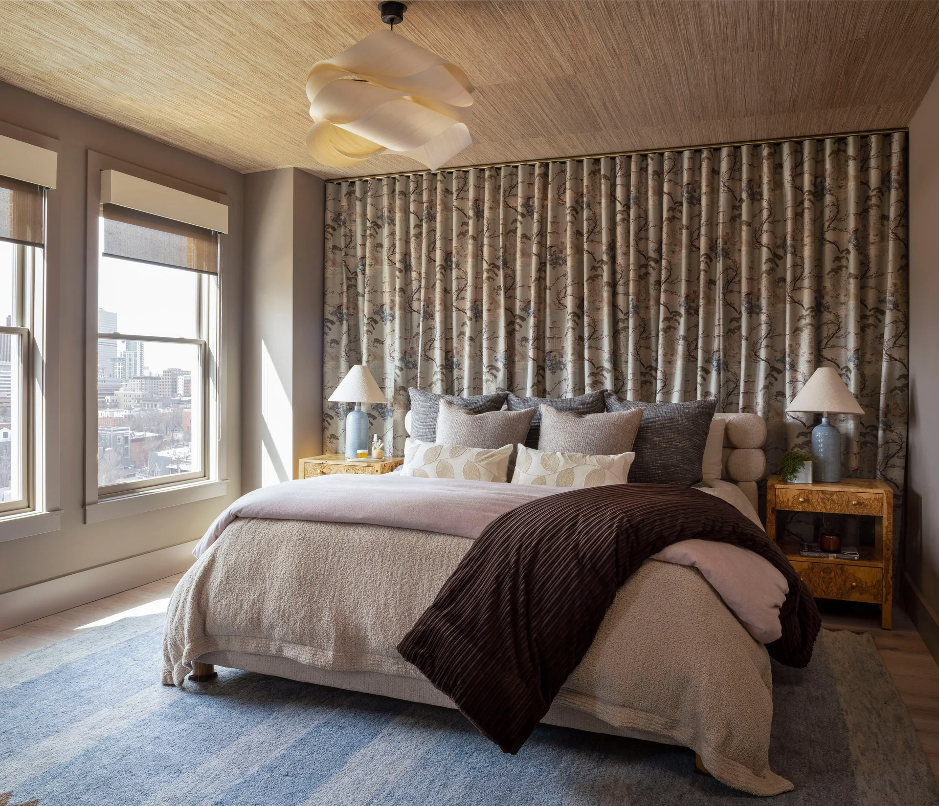 A bed with neutral linens sits against a wall of patterned floor-to-ceiling drapery, flanked by burl wood nightstands and blue table lamps.