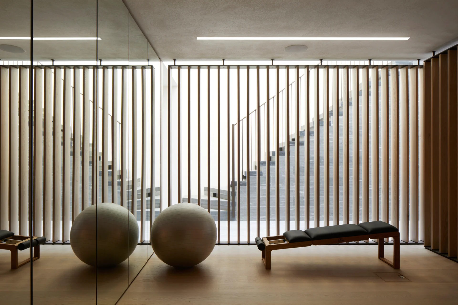 A minimalist home gym features a wooden slatted partition wall, a large floor-to-ceiling mirror, a workout bench, and a grey stability ball.