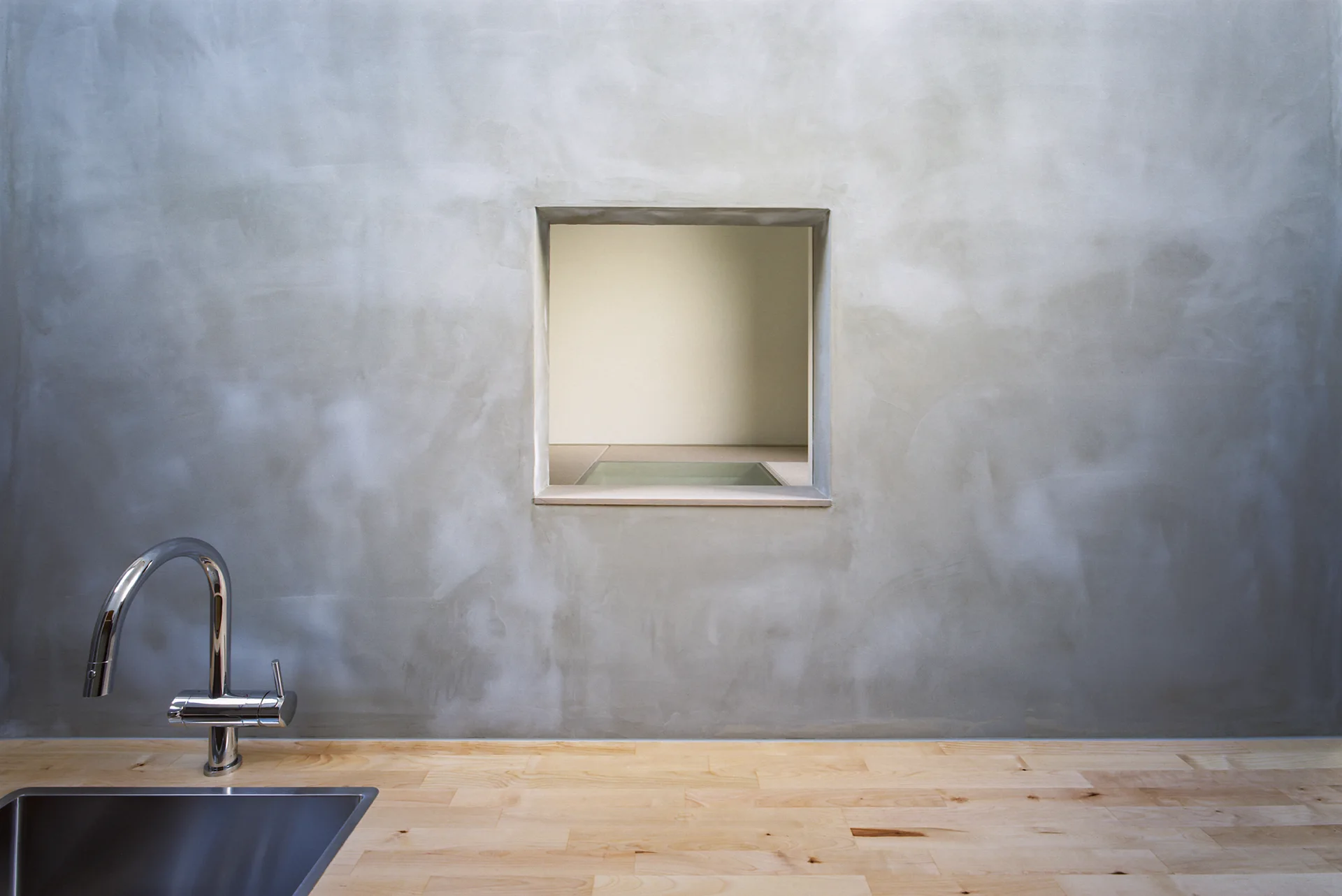 A kitchen sink with a chrome faucet set in a wood countertop below a grey concrete wall with a square interior pass-through window.