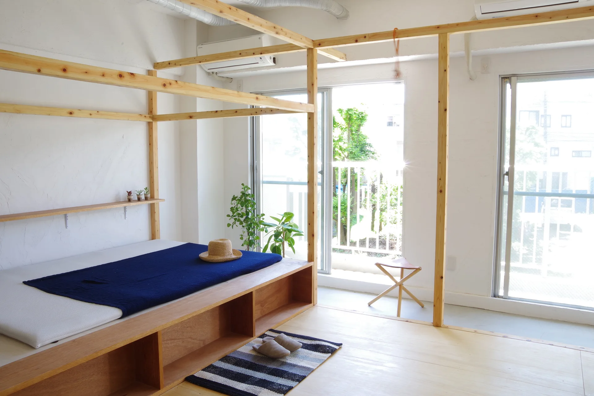 A minimalist bedroom featuring a light wood canopy bed frame with open storage cubbies below, white plaster walls, and large sliding windows.