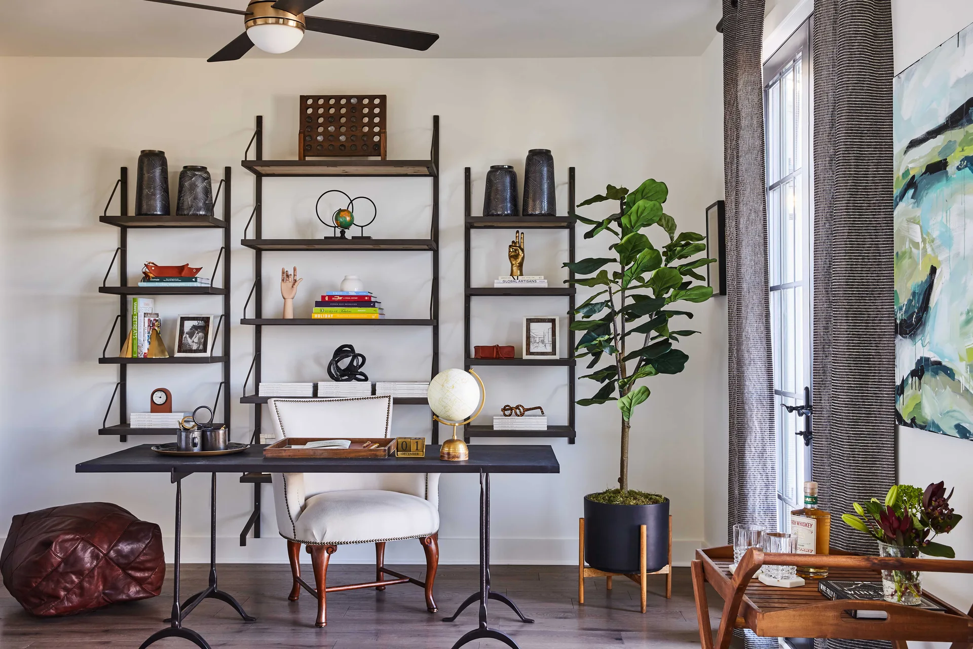 A home office features a black metal-legged desk, a white upholstered armchair, three wall-mounted industrial shelving units, and a tall potted plant.