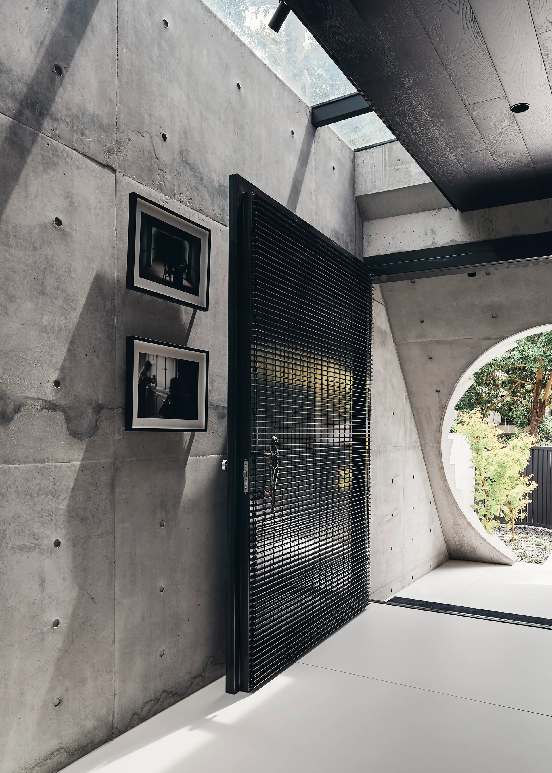 A minimalist entryway with raw concrete walls, a large black slatted door, an overhead skylight, and a circular window looking out to a garden.