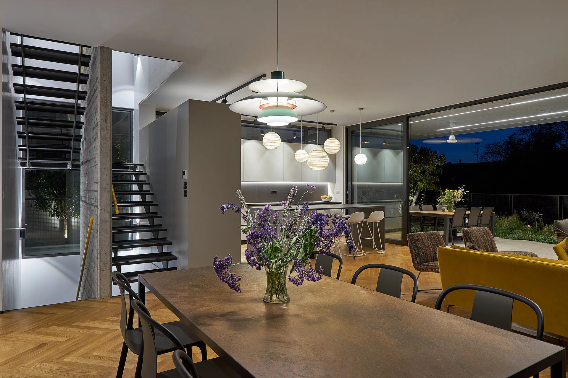 A modern dining area features a dark wood table, black chairs, a herringbone patterned floor, a metal staircase, and an open kitchen in the background.