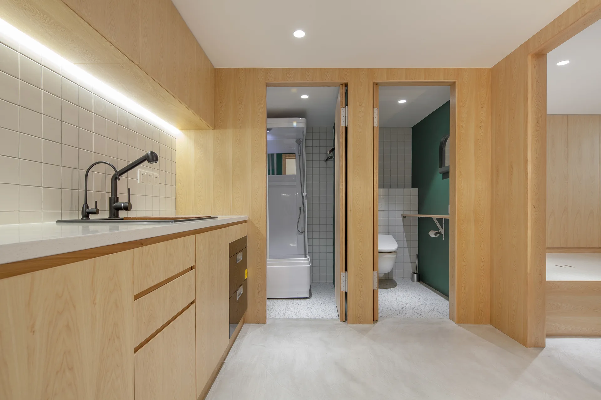A modern bathroom features a light wood vanity with a black faucet alongside two wood-framed doorways leading to separate toilet and shower stalls.